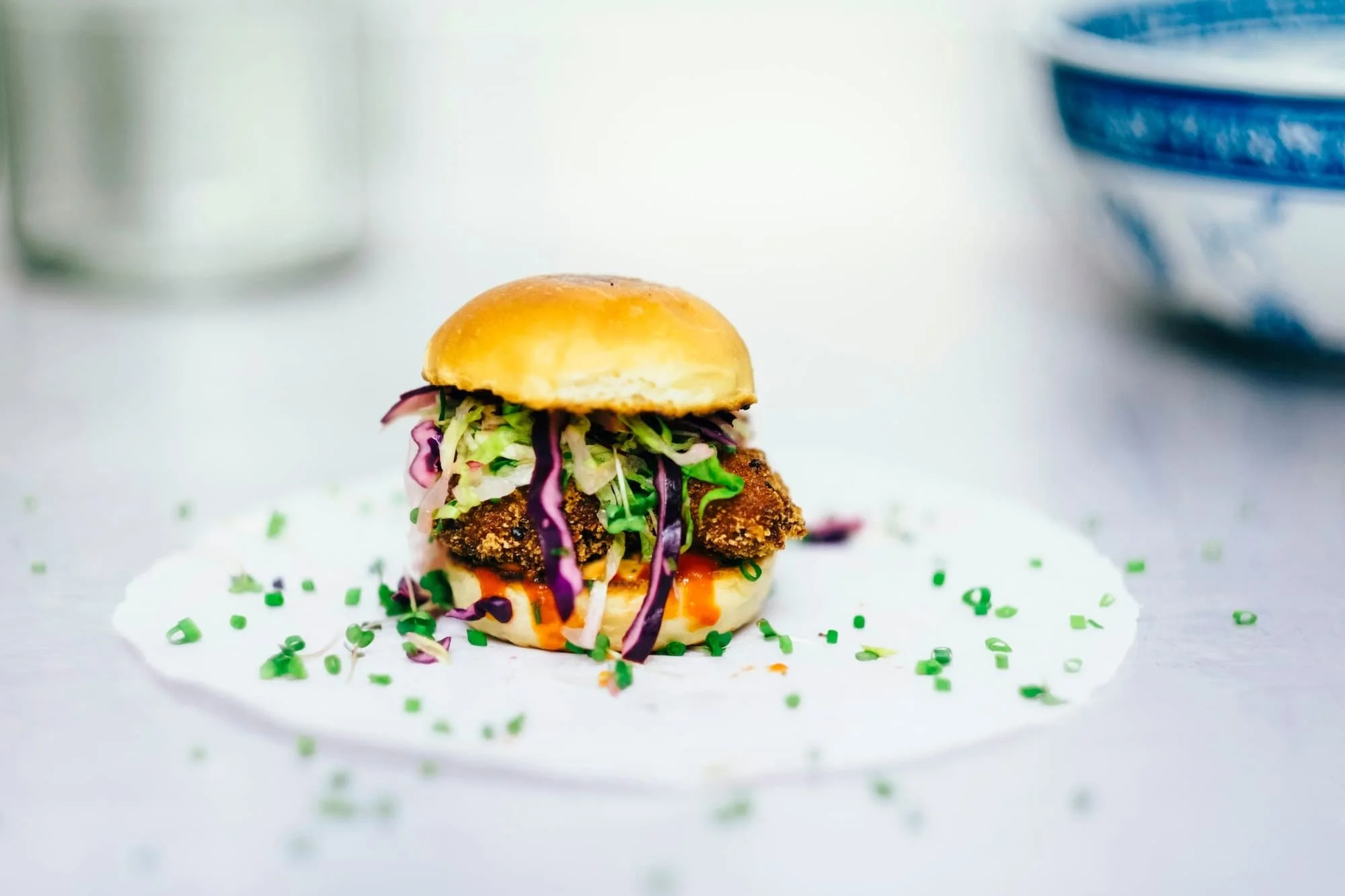 A small sandwich with fried chicken, lettuce, purple cabbage, and a bun on top, garnished with green confetti, on white paper.