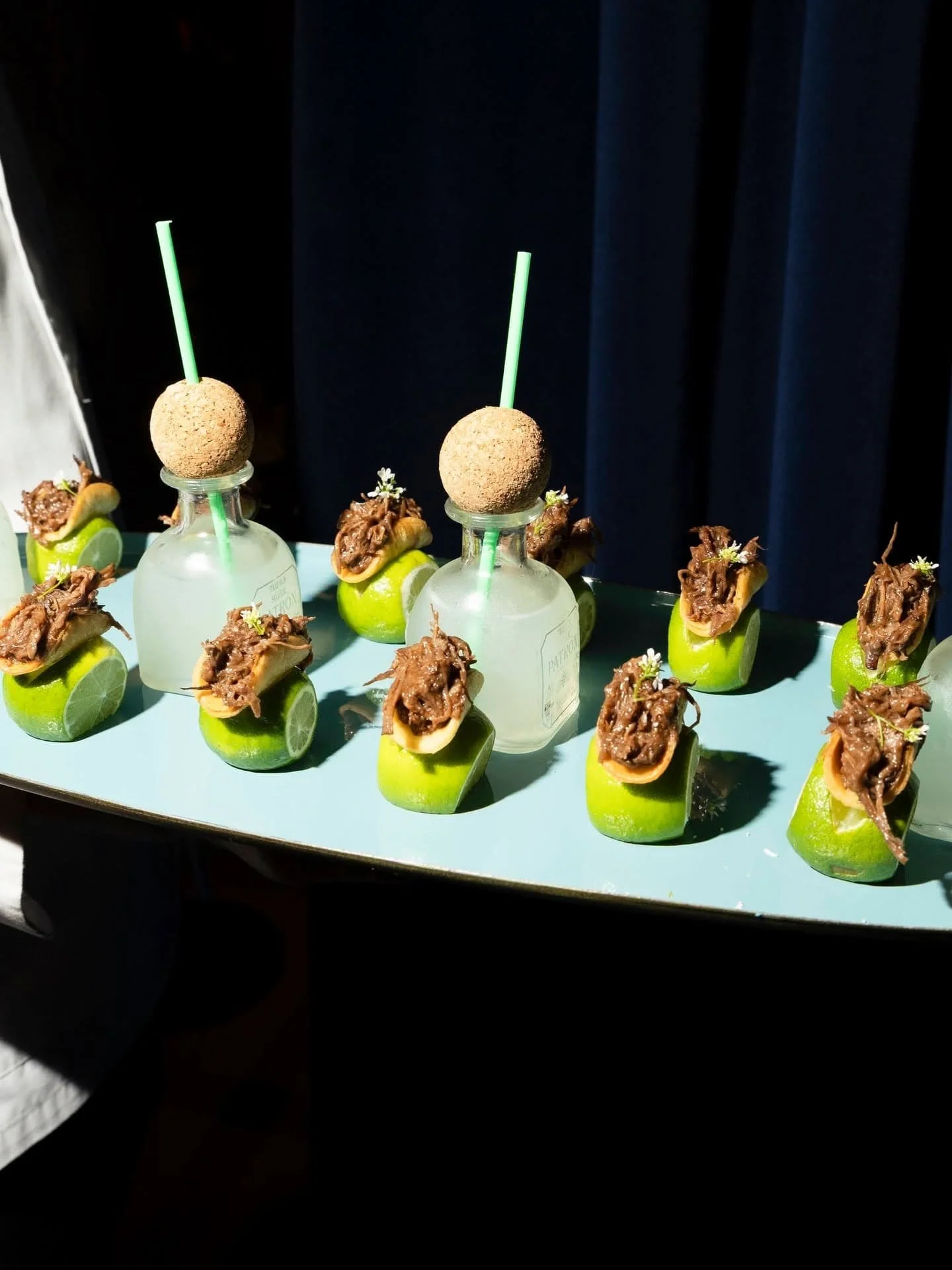Appetizer plate by Peter Callahan with halved limes topped with shredded meat and garnished with small white flowers, and two small bottles with round objects and green straws.