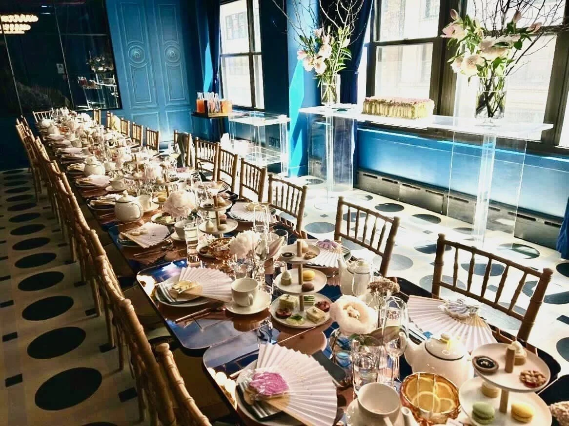 A table set for a celebration at Carton House NYC with floral centerpieces, teacups, desserts, and folded napkins in a decorated room with blue walls and large windows.