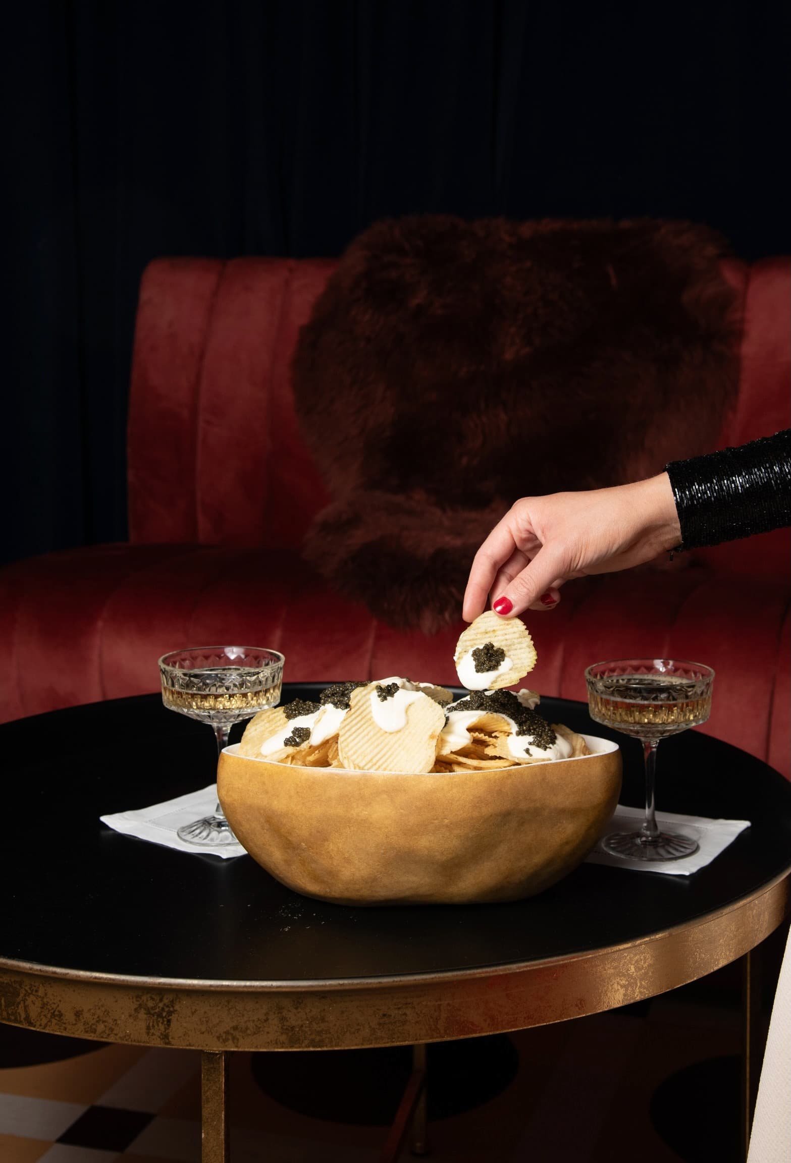 A large bowl filled with potato chips, topped with sour cream and caviar, on a black table with two glasses of champagne, in a dimly lit setting with a red cushioned bench and black curtains in the background at Carton House NYC event space