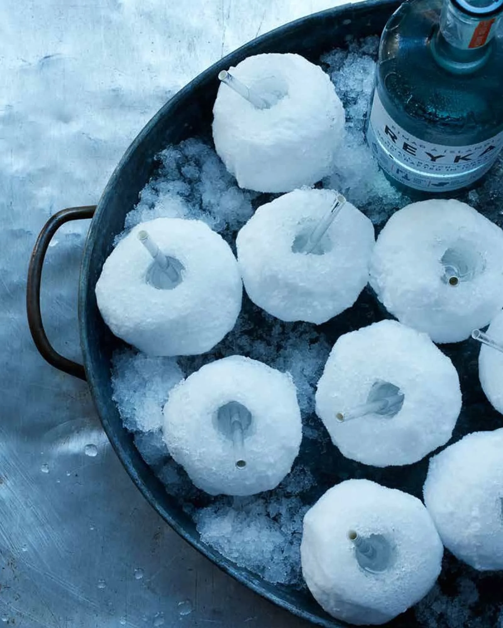 Three snow-covered ice balls with straws, a bottle of vodka, and a metal tray containing ice on a snowy surface.