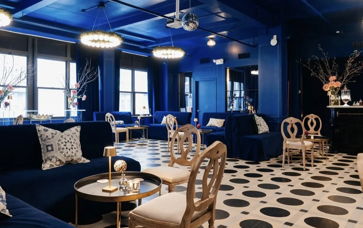 Interior of a stylish lounge area at Carton House NYC with dark blue walls and furniture, black and white patterned flooring, large windows with blue curtains, decorative tables, and floral arrangements.