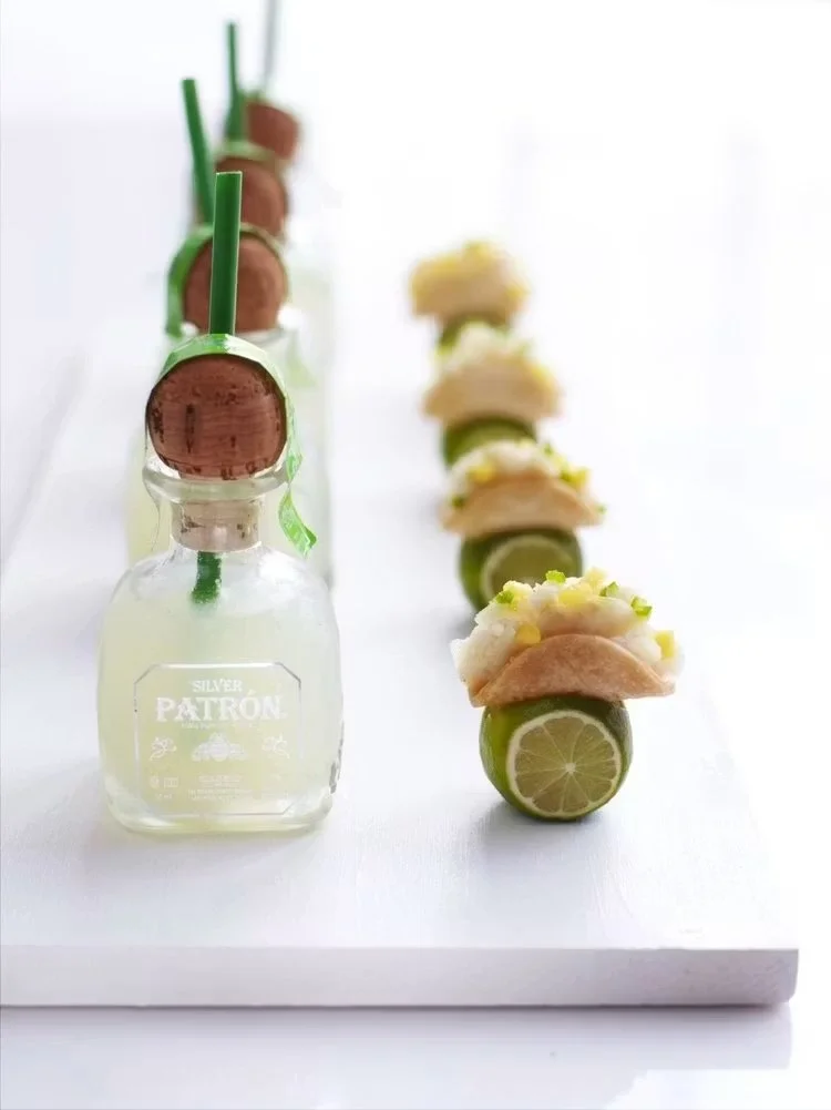 Line of shot glasses filled with tequila, each garnished with a lime slice, with a row of mini tacos on a white surface.