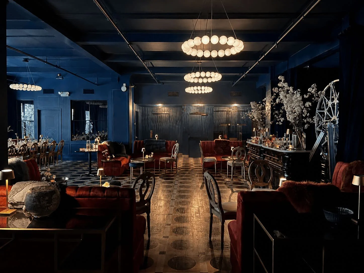 Elegant Carton House NYC event lounge with red velvet sofas, various chairs, and tables, illuminated by modern circular chandeliers, featuring dark walls and wooden flooring with patterned tile in the background.