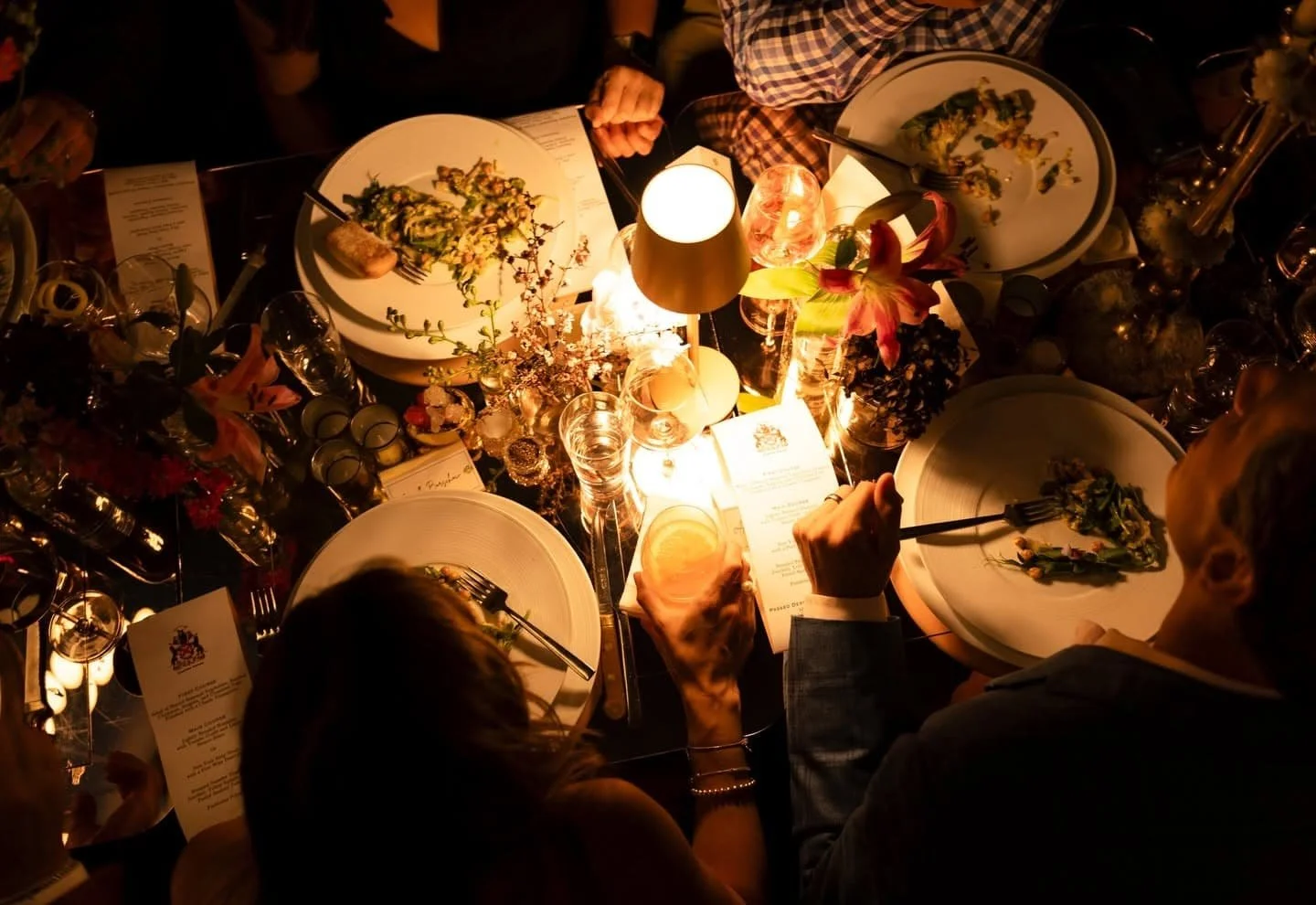 A dinner table scene at Carton House event space NYC illuminated by warm lighting, featuring plates with partially eaten food, glasses, flowers, and people serving themselves food.