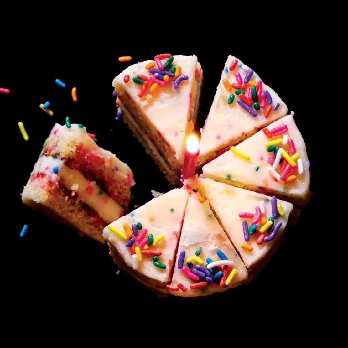 Slices of birthday cake with white frosting and colorful sprinkles, arranged in a circle with a single lit birthday candle in the center against a black background.