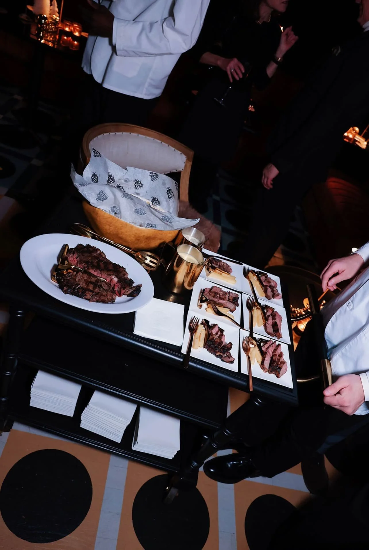 Elegant dinner party at Carton House NYC with people in formal attire, featuring a table filled with sliced steak, cheese, and small plates, set in a dimly lit room with warm lighting and candles.