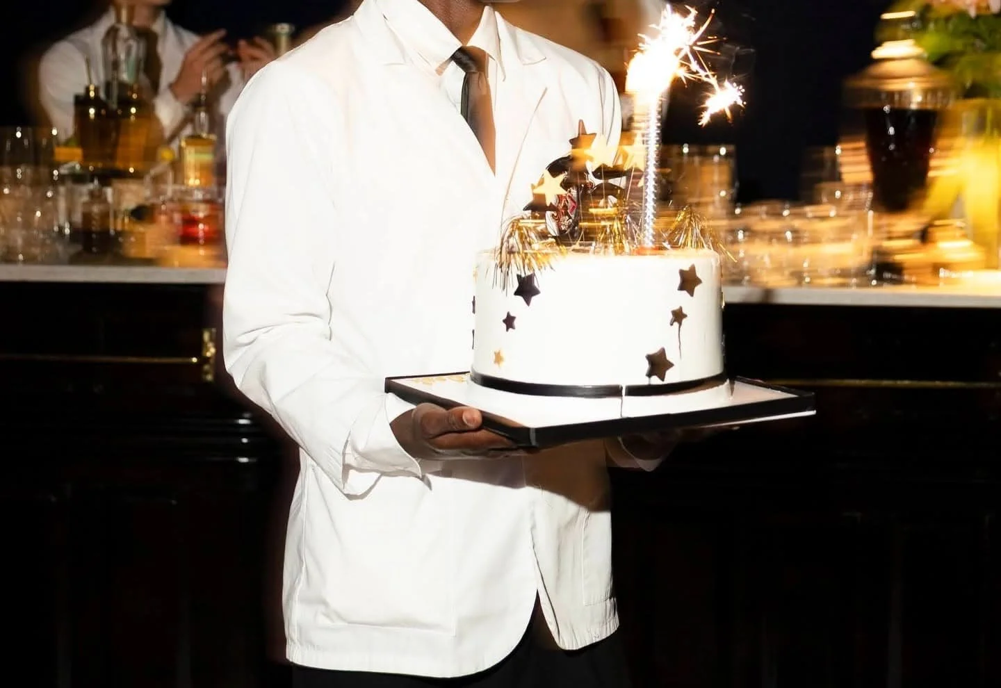 A Carton House NYC server in a white jacket and tie holding a birthday cake with sparklers and star decorations, in a festive setting at our Chealsea / NoMad private event space.
