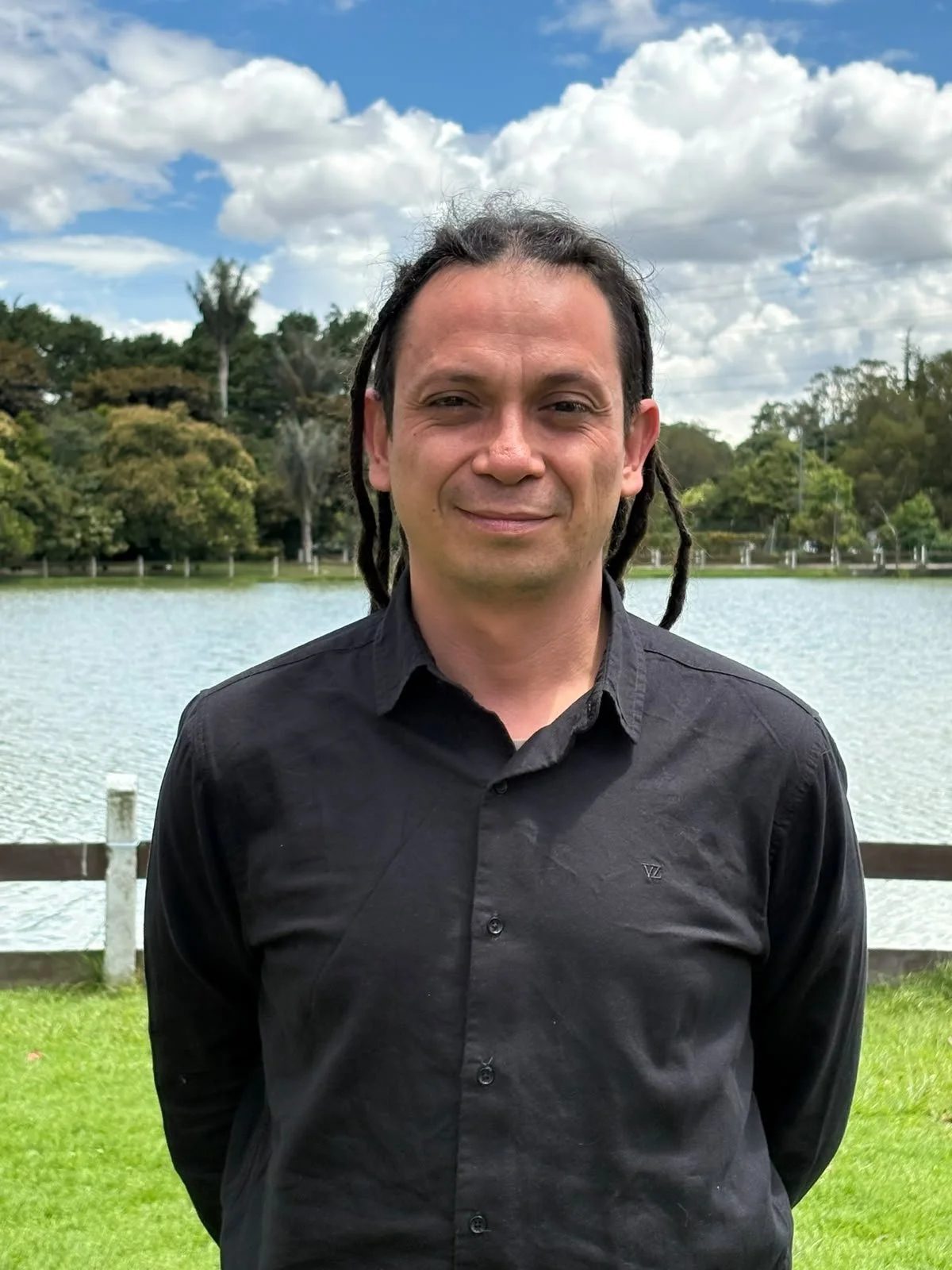 Un hombre con rastas de longitud media, que lleva una camisa negra, está de pie al aire libre cerca de un lago rodeado de árboles.
