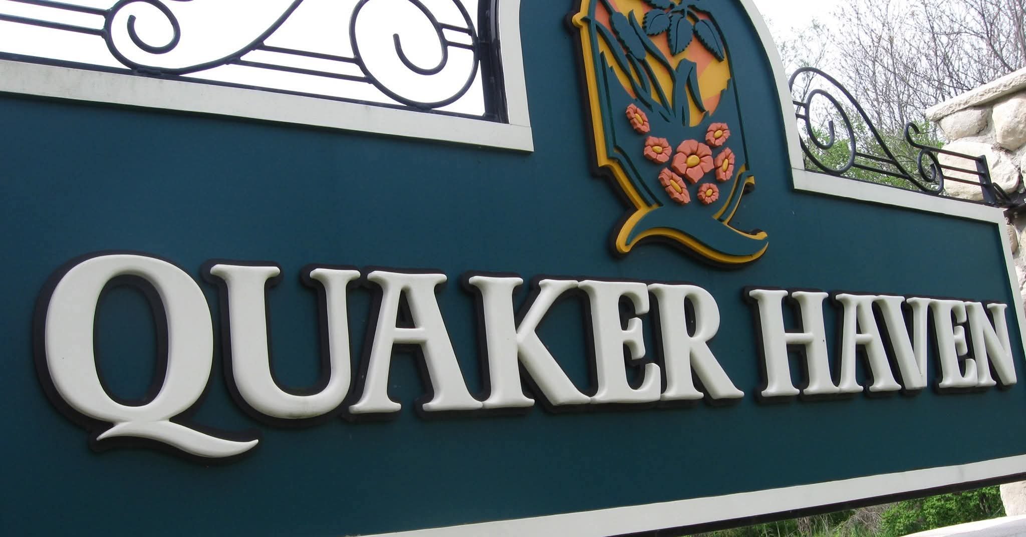 Sign with the name 'Quaker Haven' and a logo featuring a sunrise, leaves, and pink flowers, with metal decorative railings on a blue background.