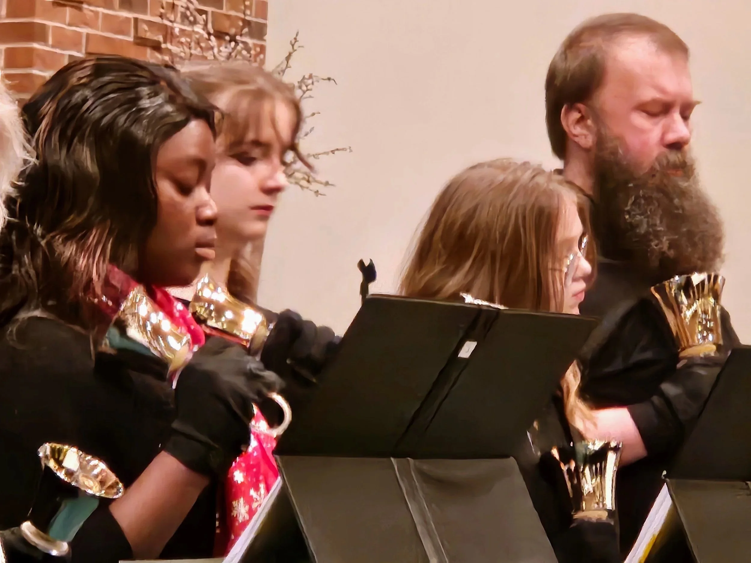 A group of people, including women and a man, performing music together, wearing festive clothing with shiny gold accents, and playing musical instruments with sheet music in front of them.