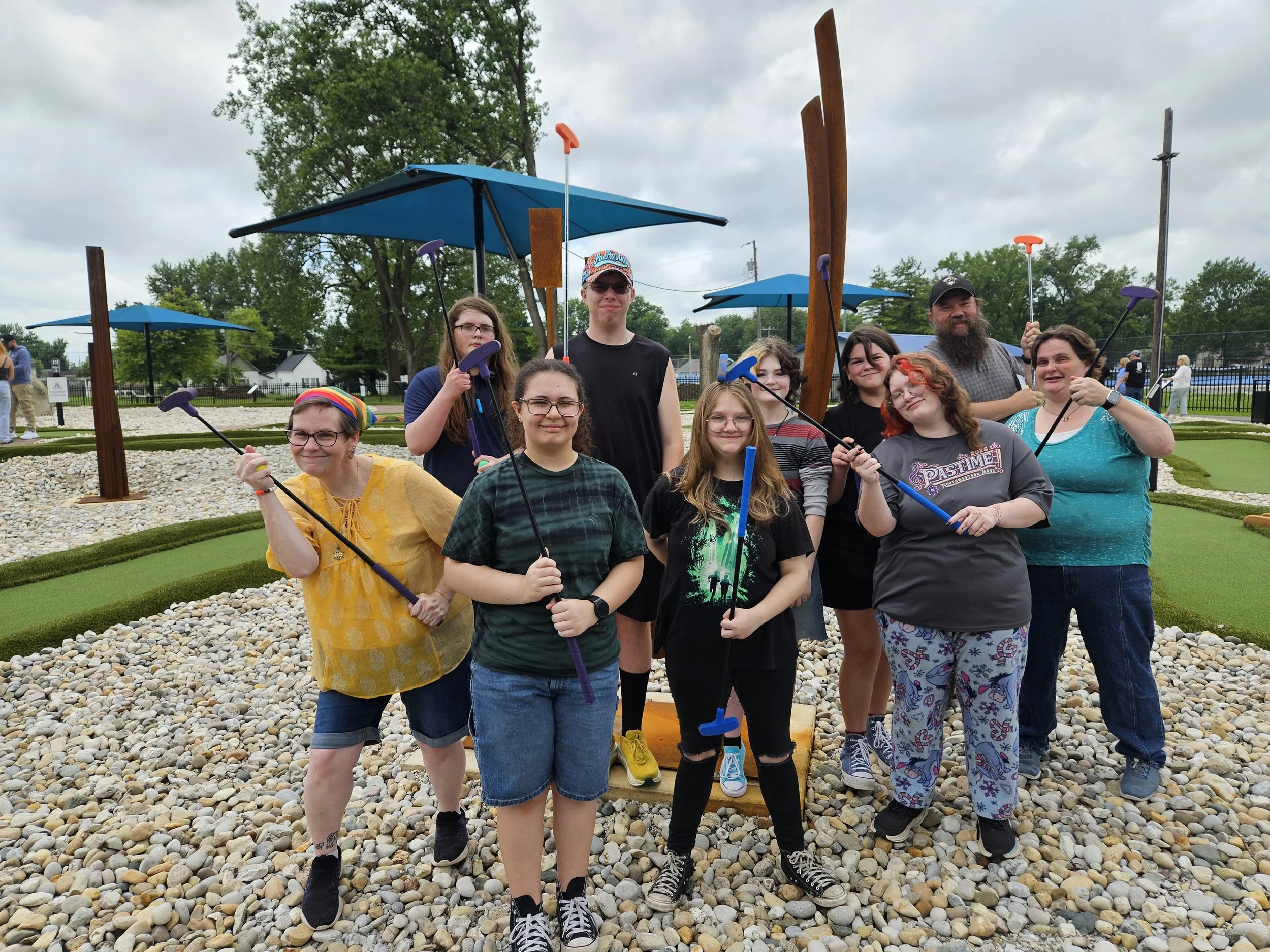 Group of nine people posing with putters on a mini-golf course with rock landscaping, blue umbrellas, trees, a fence, and cloudy skies in the background.