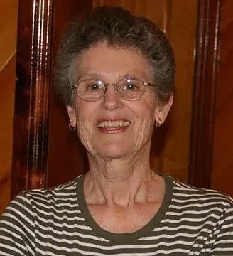 An elderly woman with short gray hair, wearing glasses and a striped shirt, smiling in front of a wooden background.