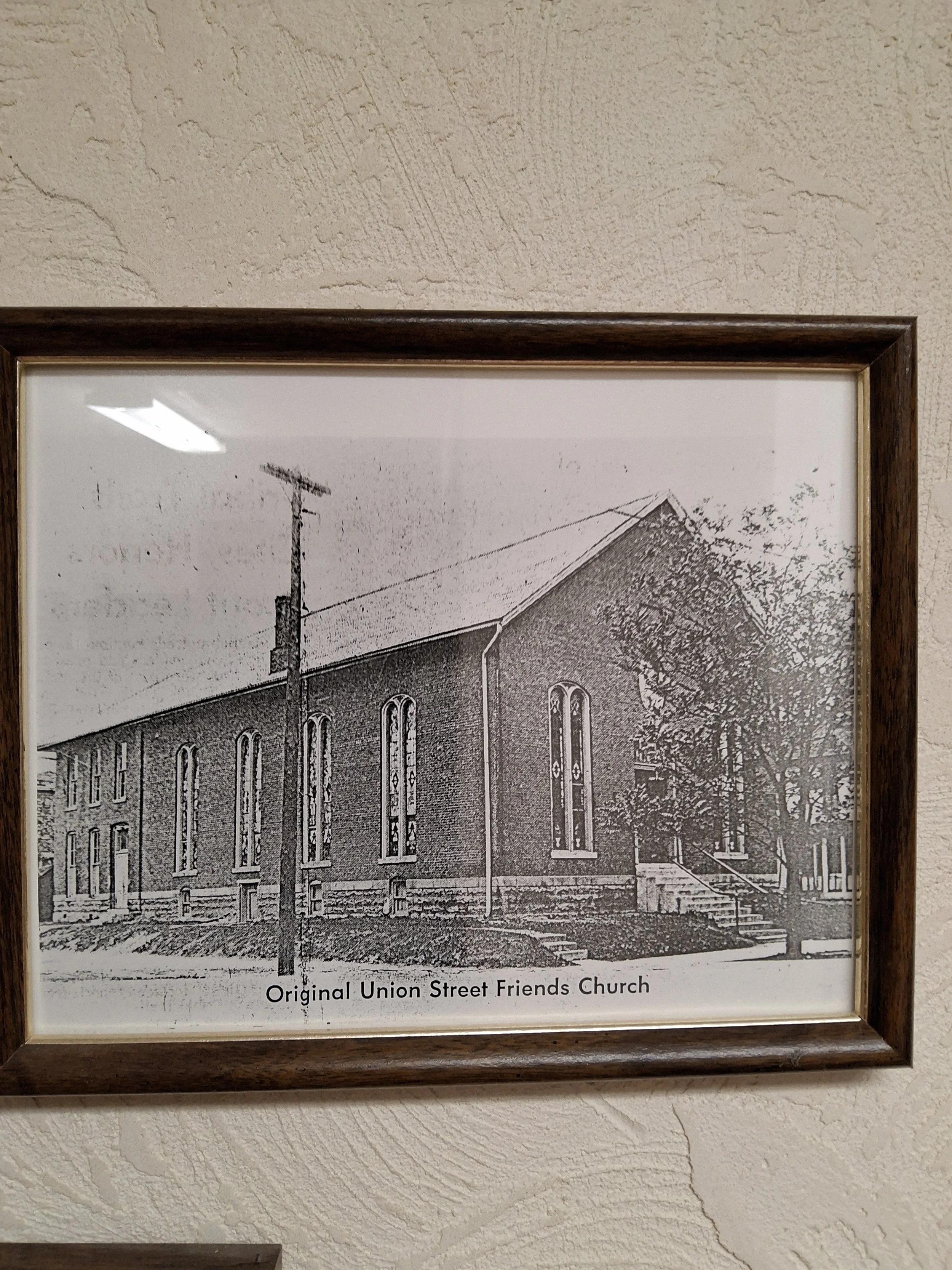 Photograph of a framed historical black-and-white illustration of the Original Union Street Friends Church. The drawing shows the church building with arched windows, a chimney, and a large door, with a street lamp and trees nearby. The frame is wooden, and a caption at the bottom reads 'Original Union Street Friends Church'.
