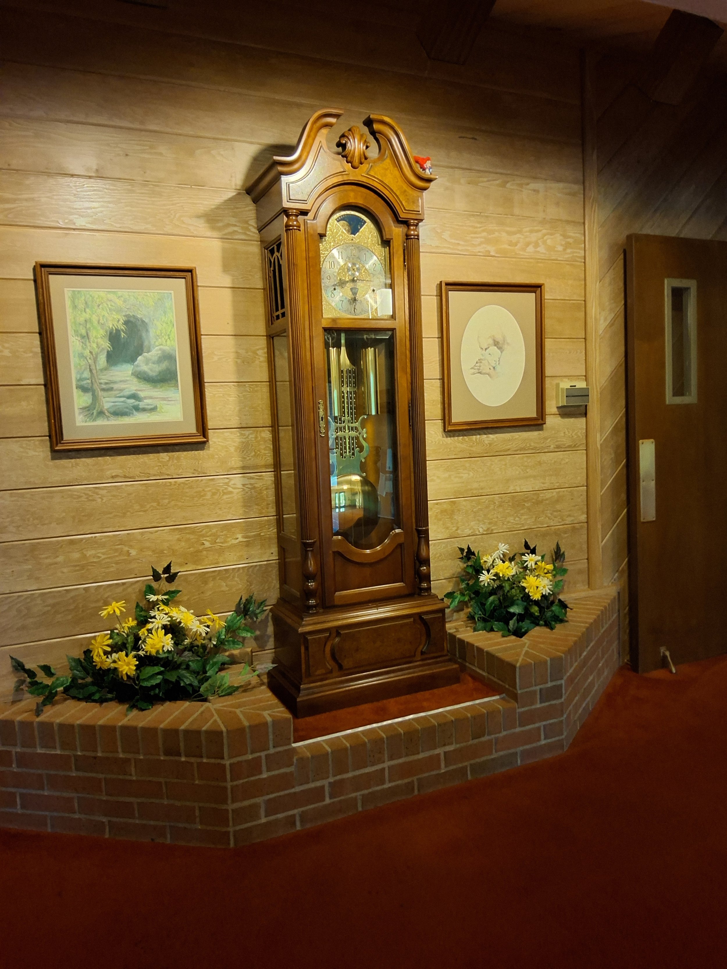 A wooden grandfather clock stands on a brick platform in a room with wood-paneled walls, surrounded by yellow flowers. Two framed pictures hang on the wall behind the clock.
