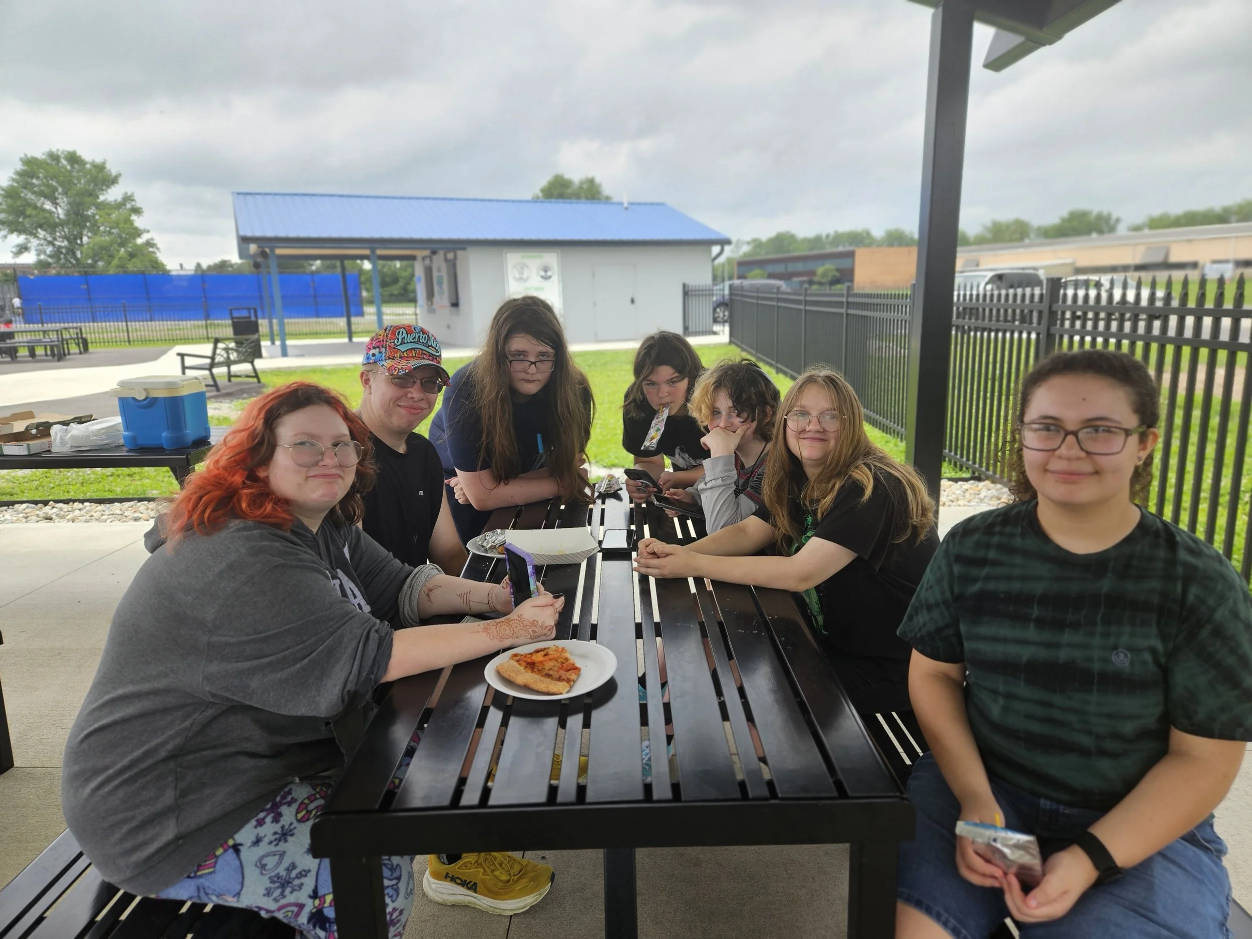 A group of people sitting at a picnic table outdoors, enjoying pizza, with a playground and cloudy sky in the background.