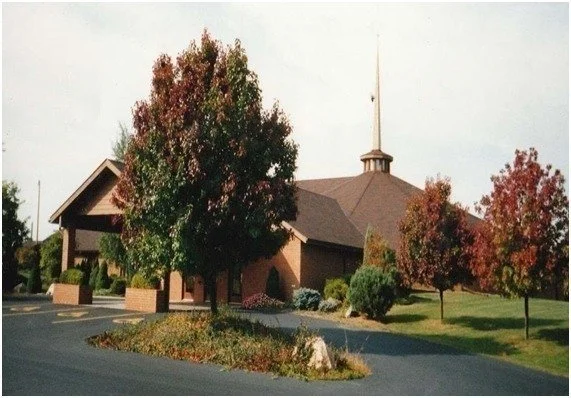 A church with a steeple surrounded by trees with autumn foliage, a parking lot, and landscaped bushes.