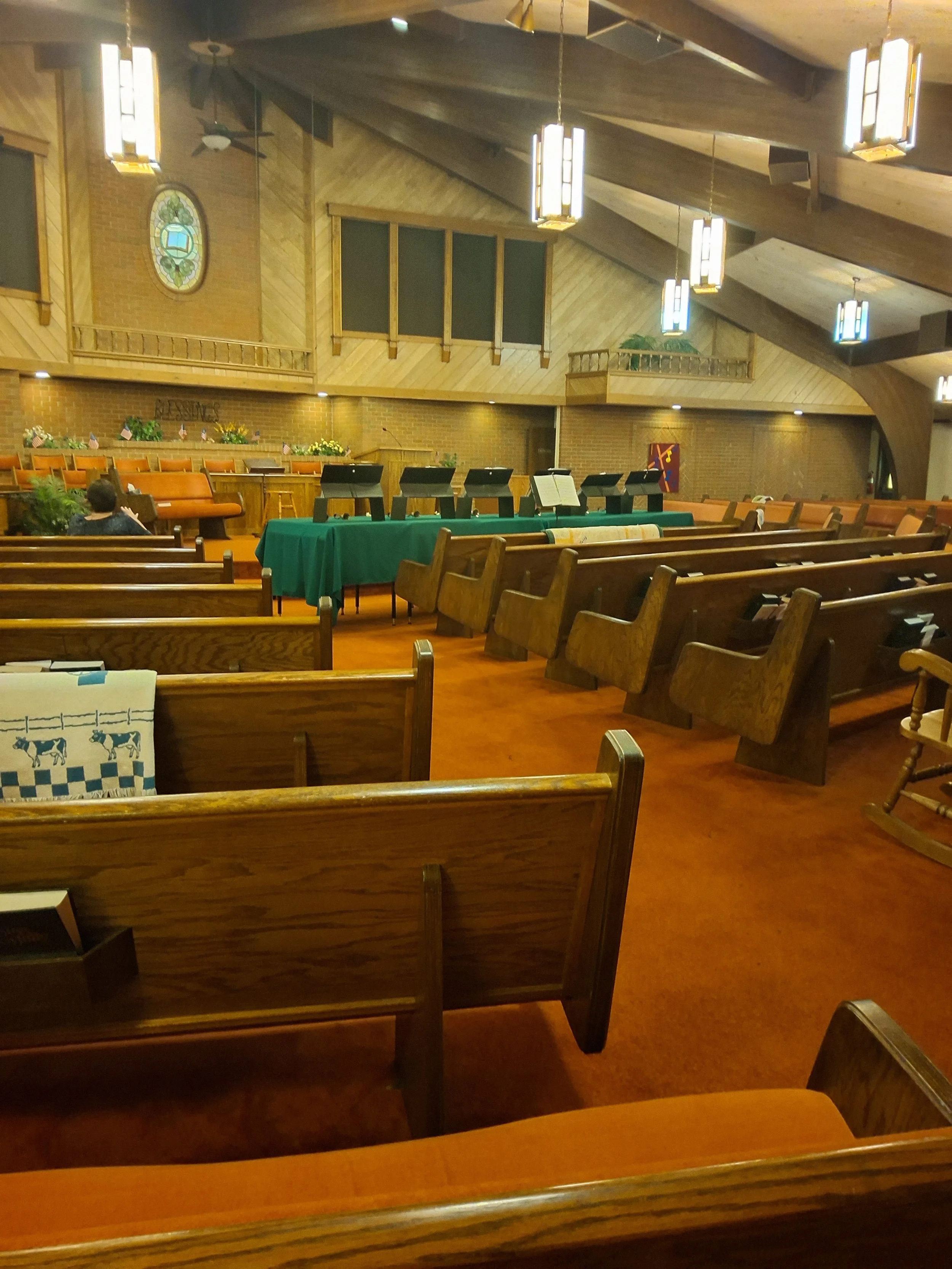 The interior of a church with wooden pews, a green tablecloth on a table with music stands, hanging lights, brick and wood walls, a stained glass window, and a person sitting in a pew.