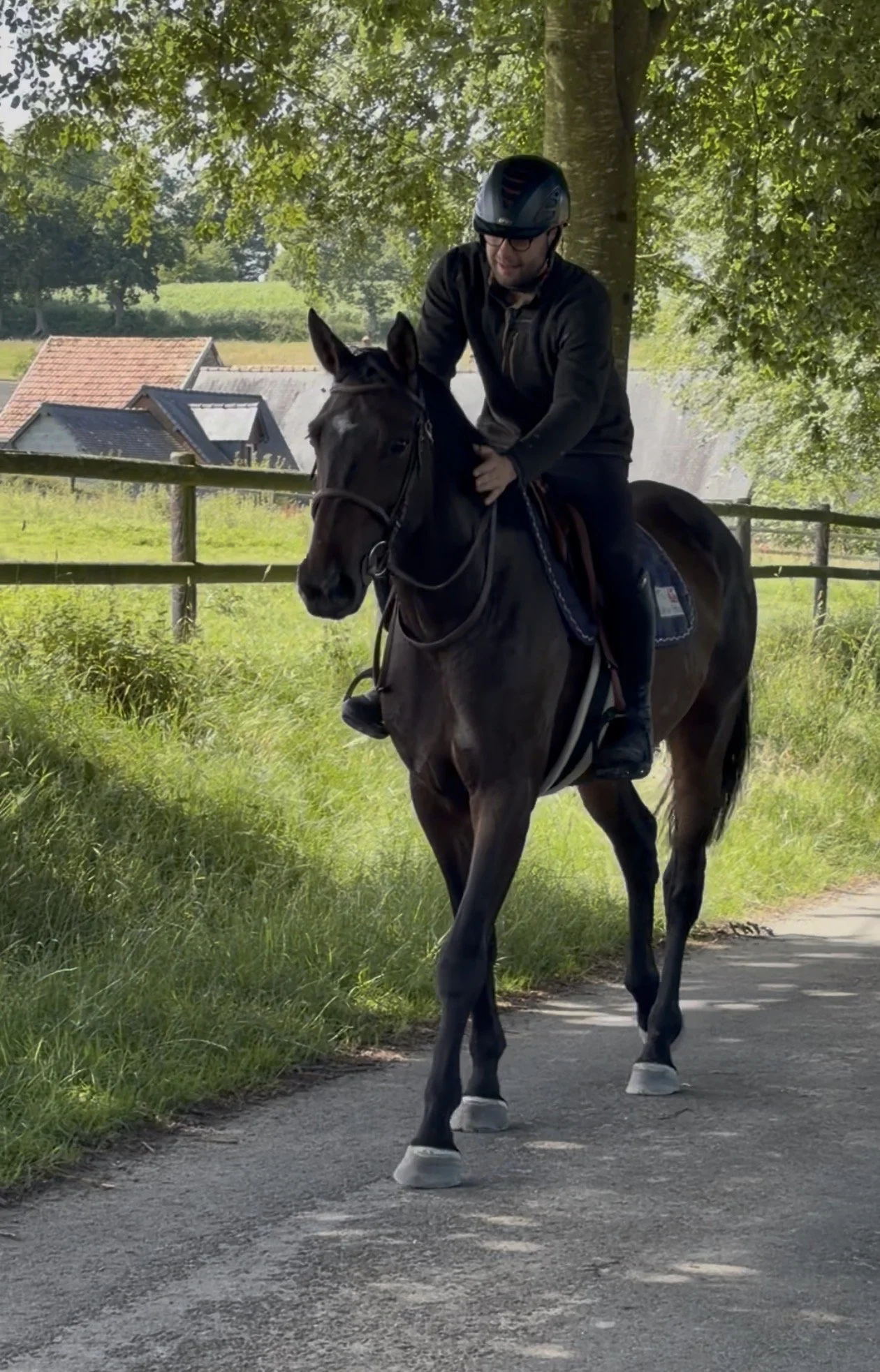 Homme à cheval portant un casque, chevauchant sur un chemin dans un environnement rural avec des arbres, un panneau de clôture en bois et des maisons en arrière-plan.