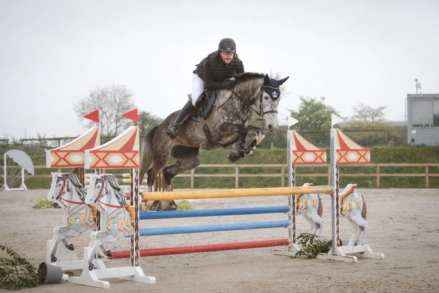 Un cavalier en noir et blanc chevauche un cheval gris clair en saut d'obstacles lors d'une compétition équestre, avec un décor de chevaux en bois decoratifs et un ciel nuageux.
