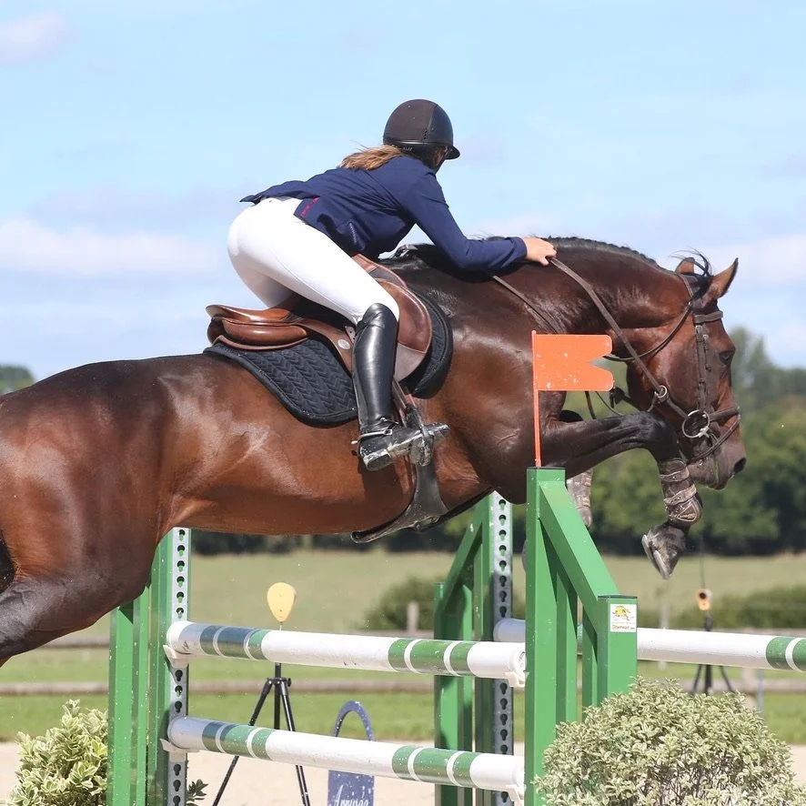 Une cavalière en tenue de concours équestre sautant une obstacle avec son cheval dans un champ.