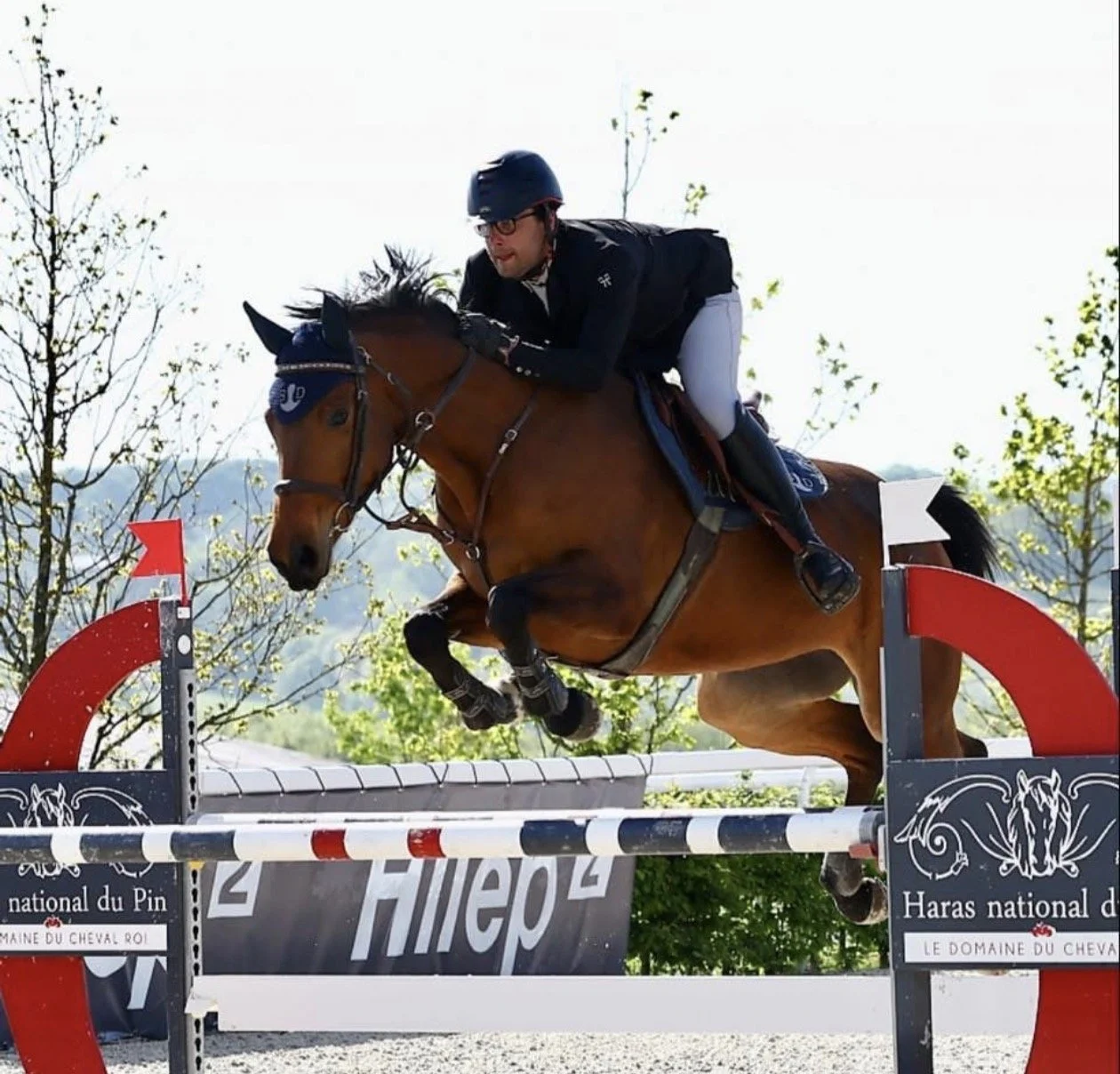 Un cavalier en tenue noire et blanche, portant un casque et des lunettes, saute une barrière lors d'une compétition équestre en extérieur