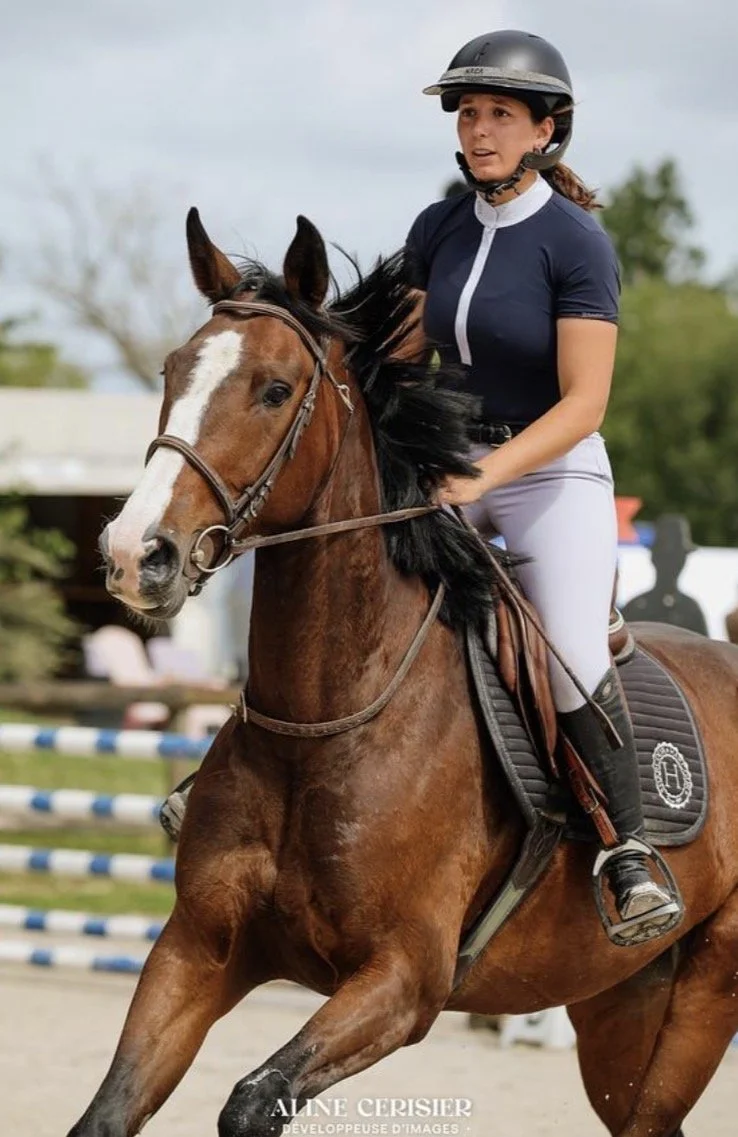 Une femme en équipement d'équitation, portant un casque noir, chevauche un cheval alezan avec une tache blanche sur le front, lors d'une compétition équestre en plein air.