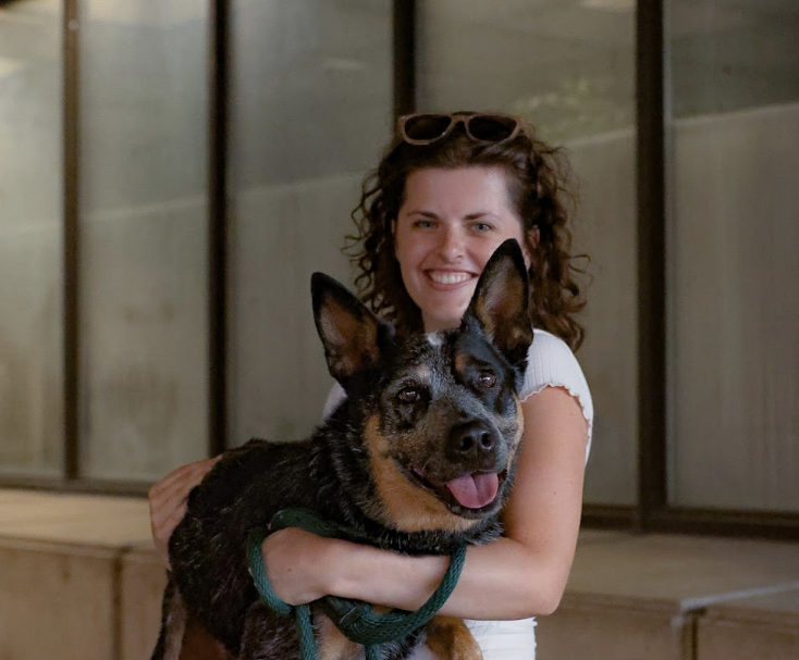 A woman with curly hair wearing sunglasses on her head, smiling and hugging a black and tan dog with a happy expression, in an indoor setting with glass doors in the background.