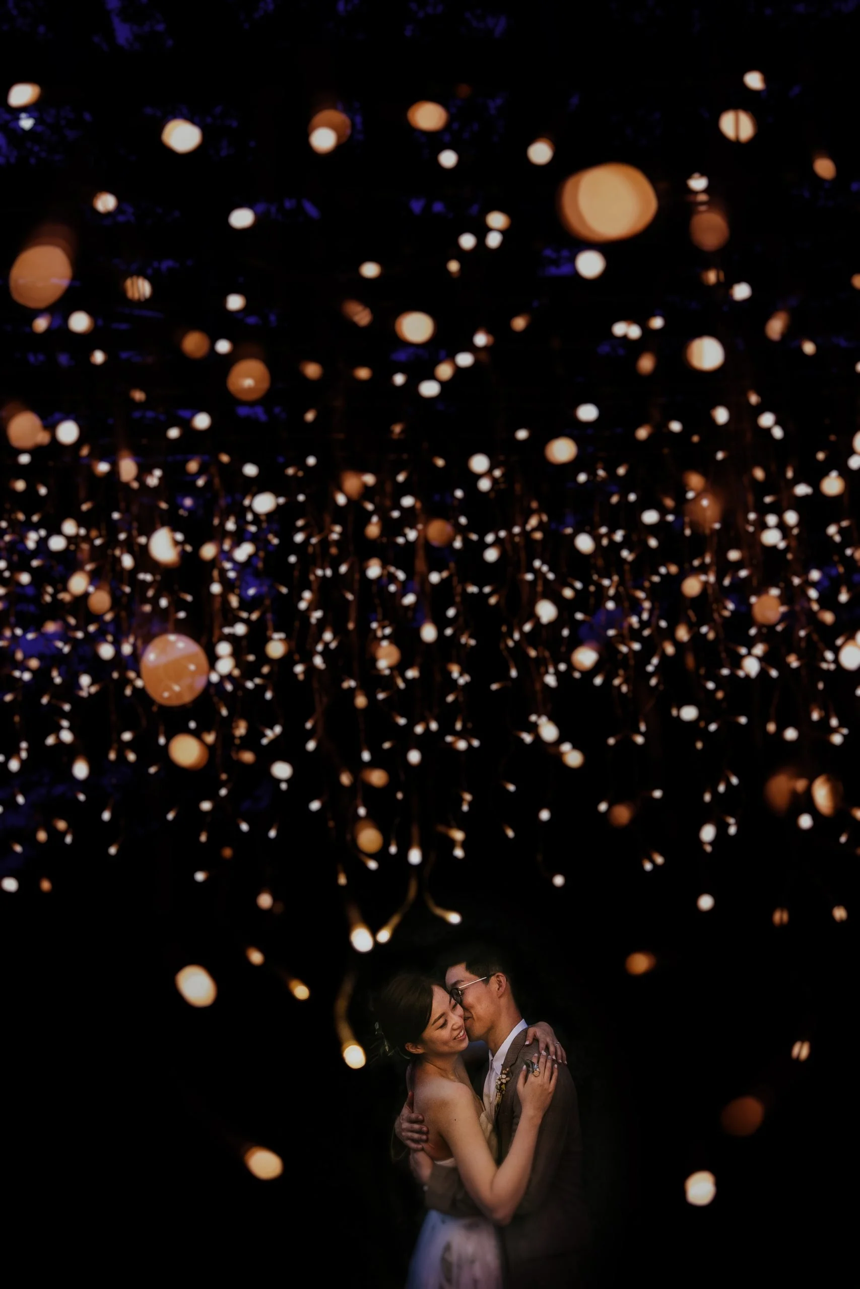 A couple dancing closely under a canopy of hanging string lights at night.