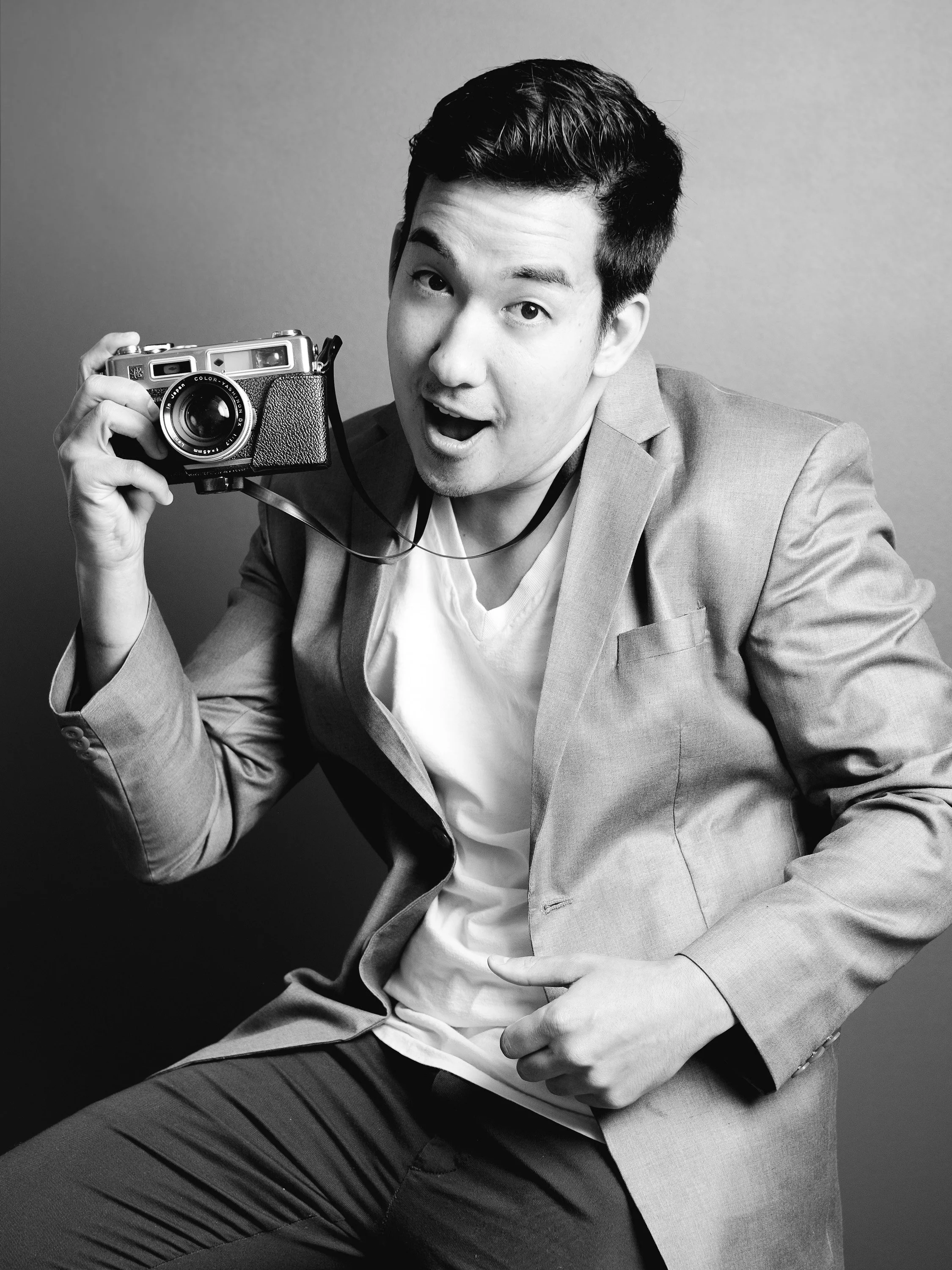 A young man in a suit making a surprised or excited facial expression while holding a vintage camera in his right hand, sitting against a plain background.