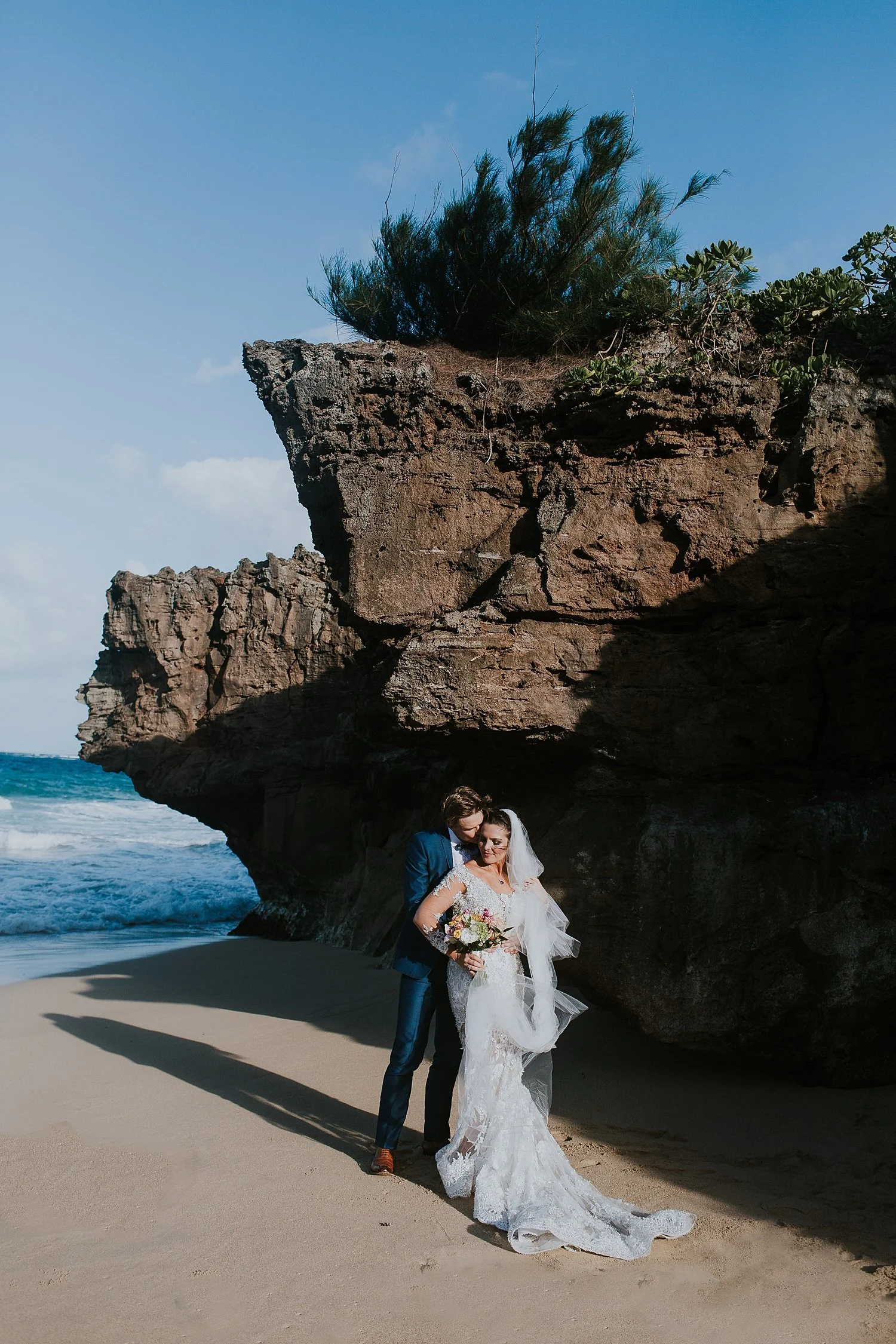 A newlywed couple in wedding attire standing on a sandy beach near a rocky cliff with green plants, ocean waves, and a blue sky in the background.