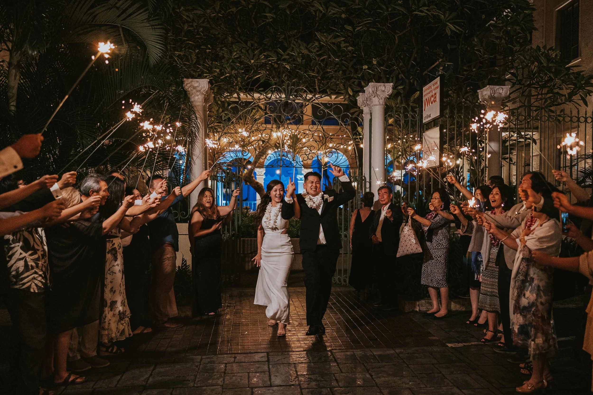 A newlywed couple walking through a crowd holding sparklers at night outside a building with ornate gates and trees.