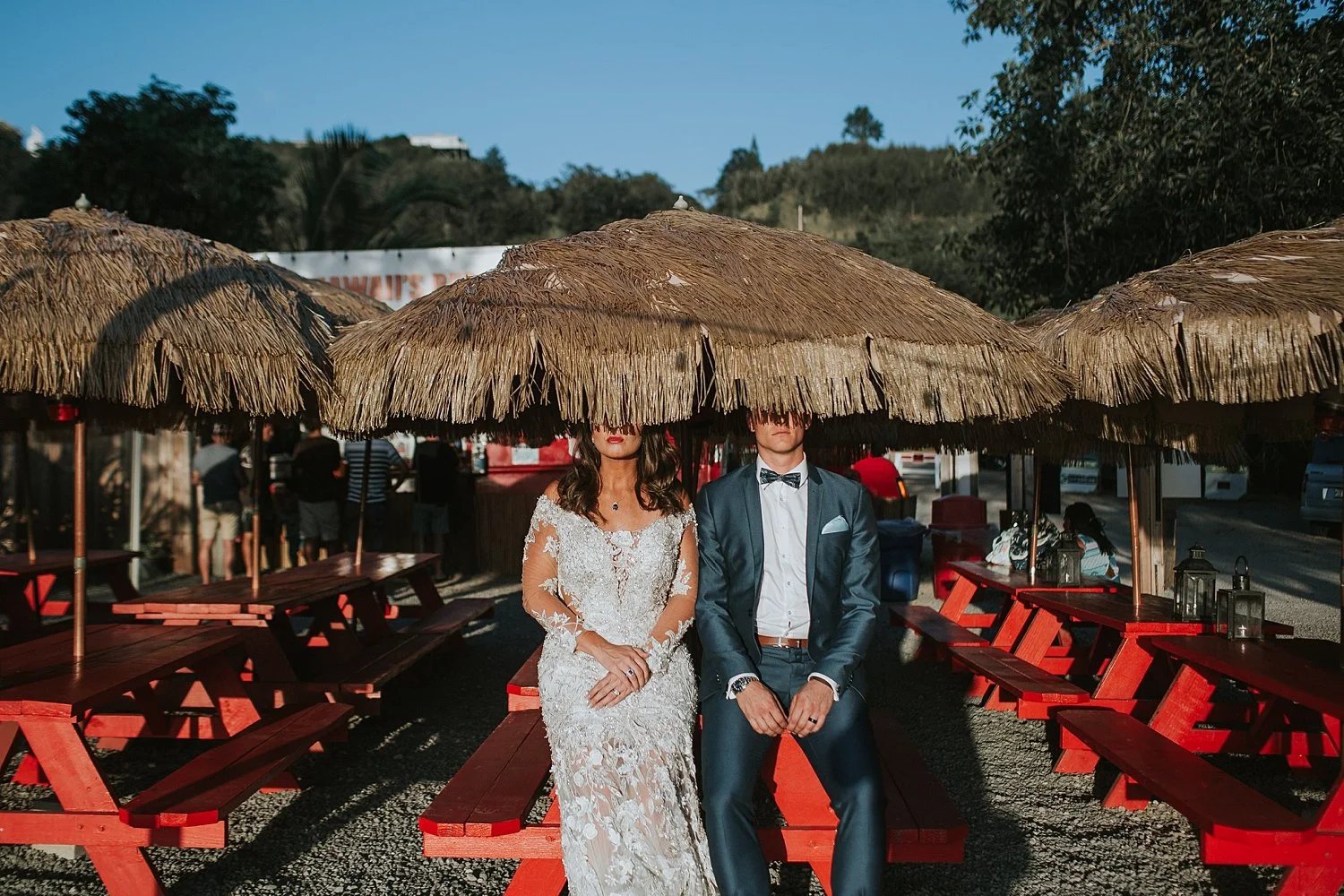 A bride and groom sitting under a large straw umbrella on red picnic benches at an outdoor event.