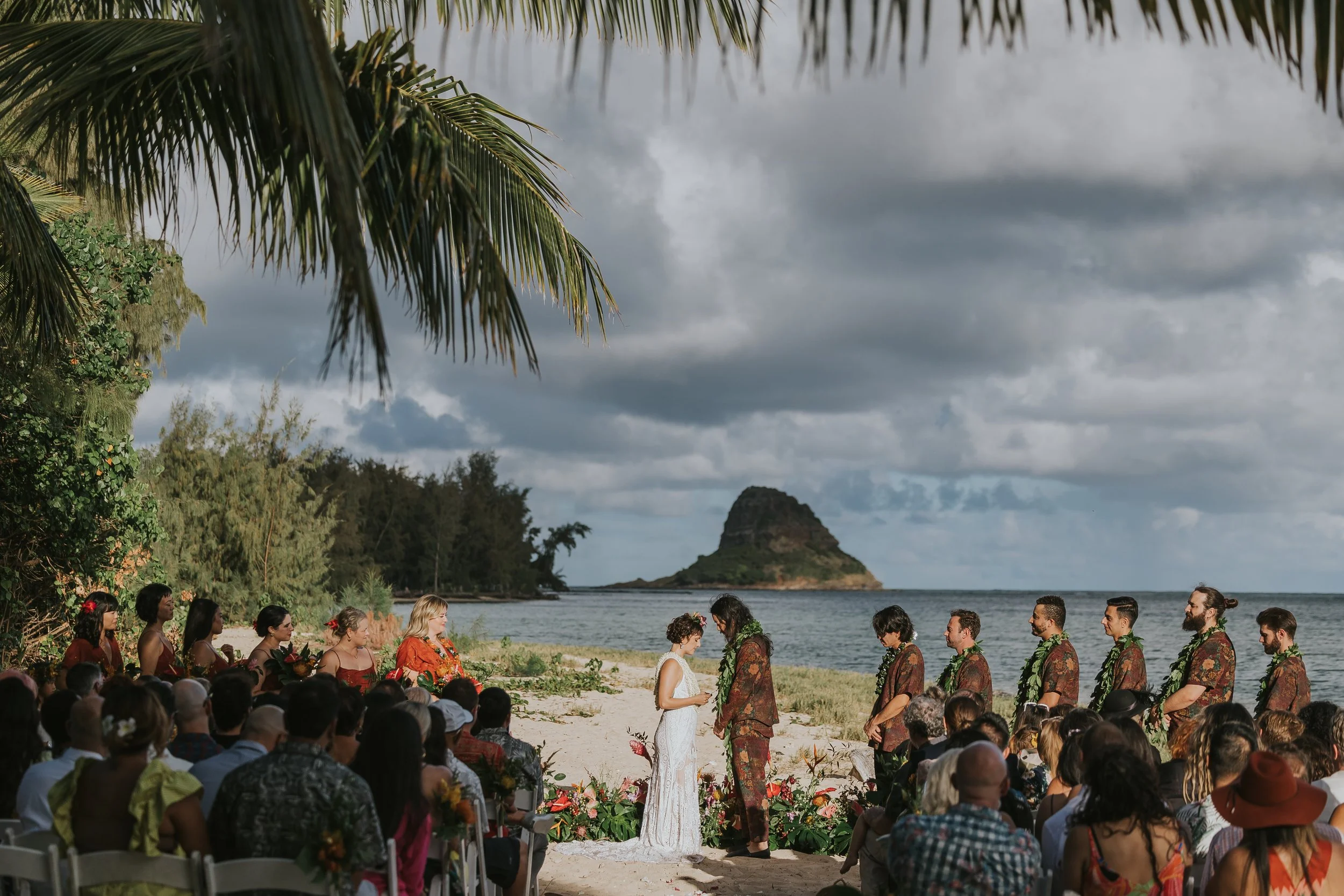 A wedding ceremony on a beach with a group of people, tropical trees, and an island in the background under cloudy skies.