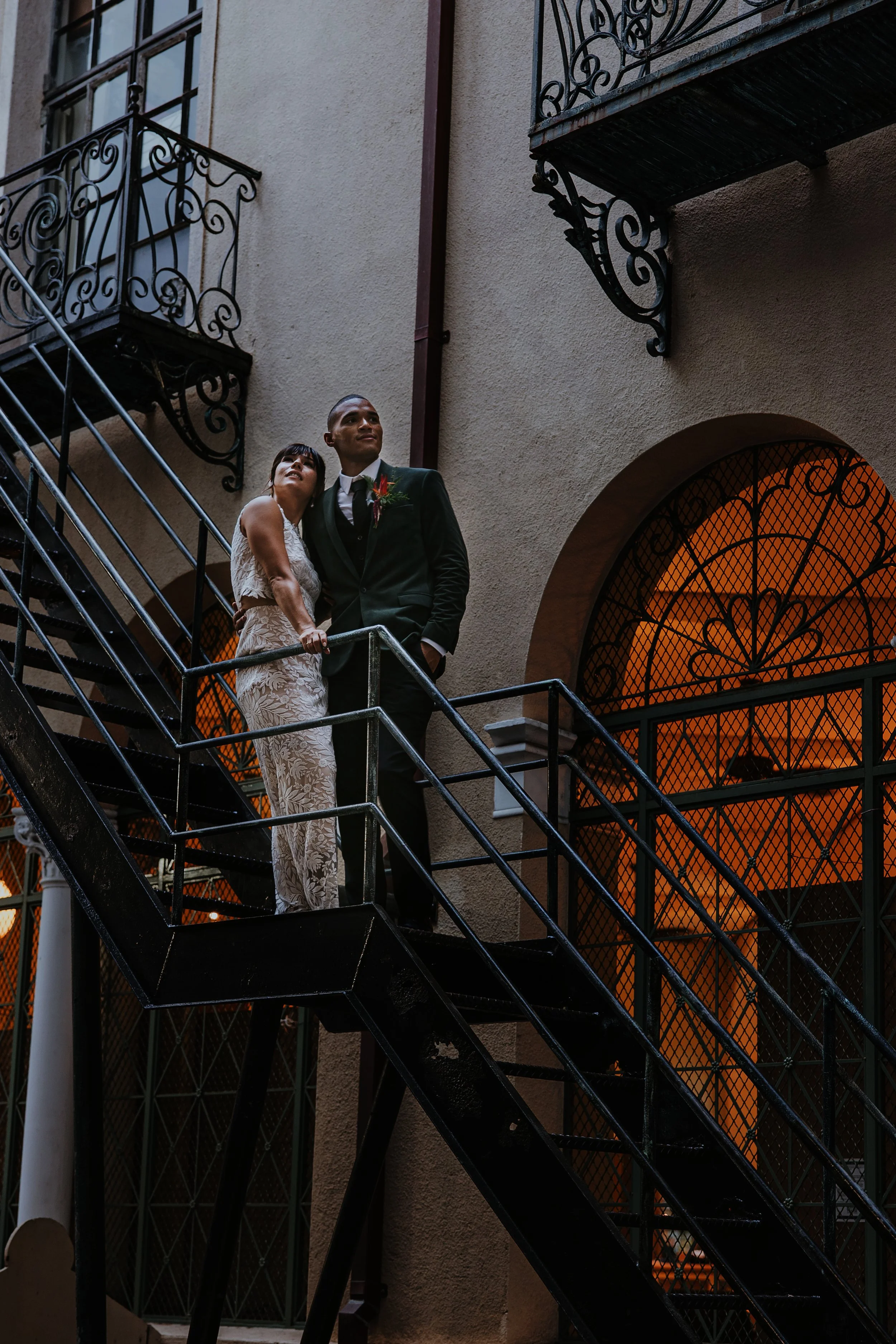 A bride and groom standing on an outdoor staircase with ornate black metal railings, in front of a building with large arched windows with warm orange interior lighting.
