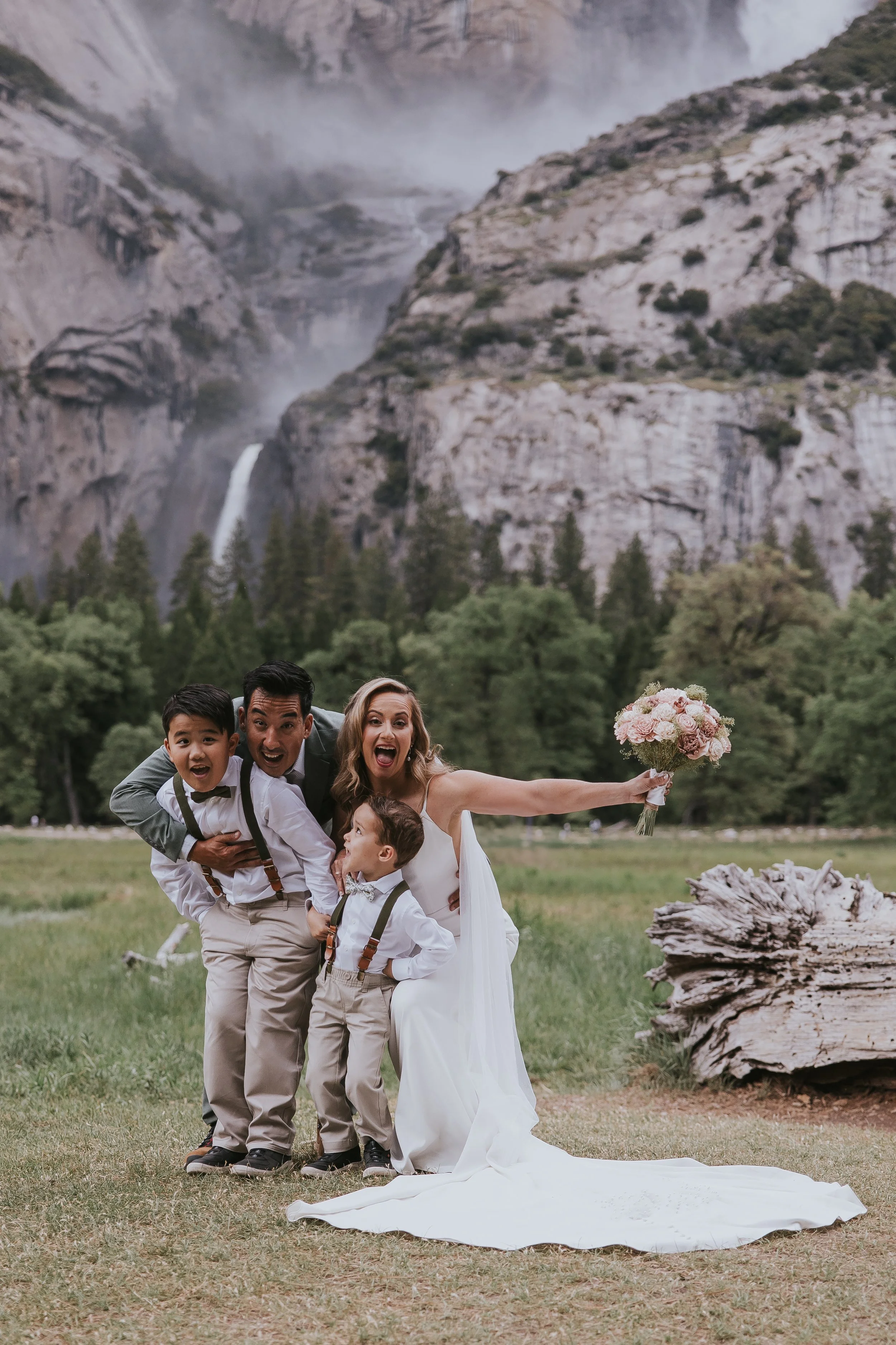 A joyful wedding party consisting of a bride, three young boys, and a man posing outdoors on a grassy field with mountains, a waterfall, and trees in the background. The bride holds a bouquet of pink flowers, and all are smiling and enjoying the moment.