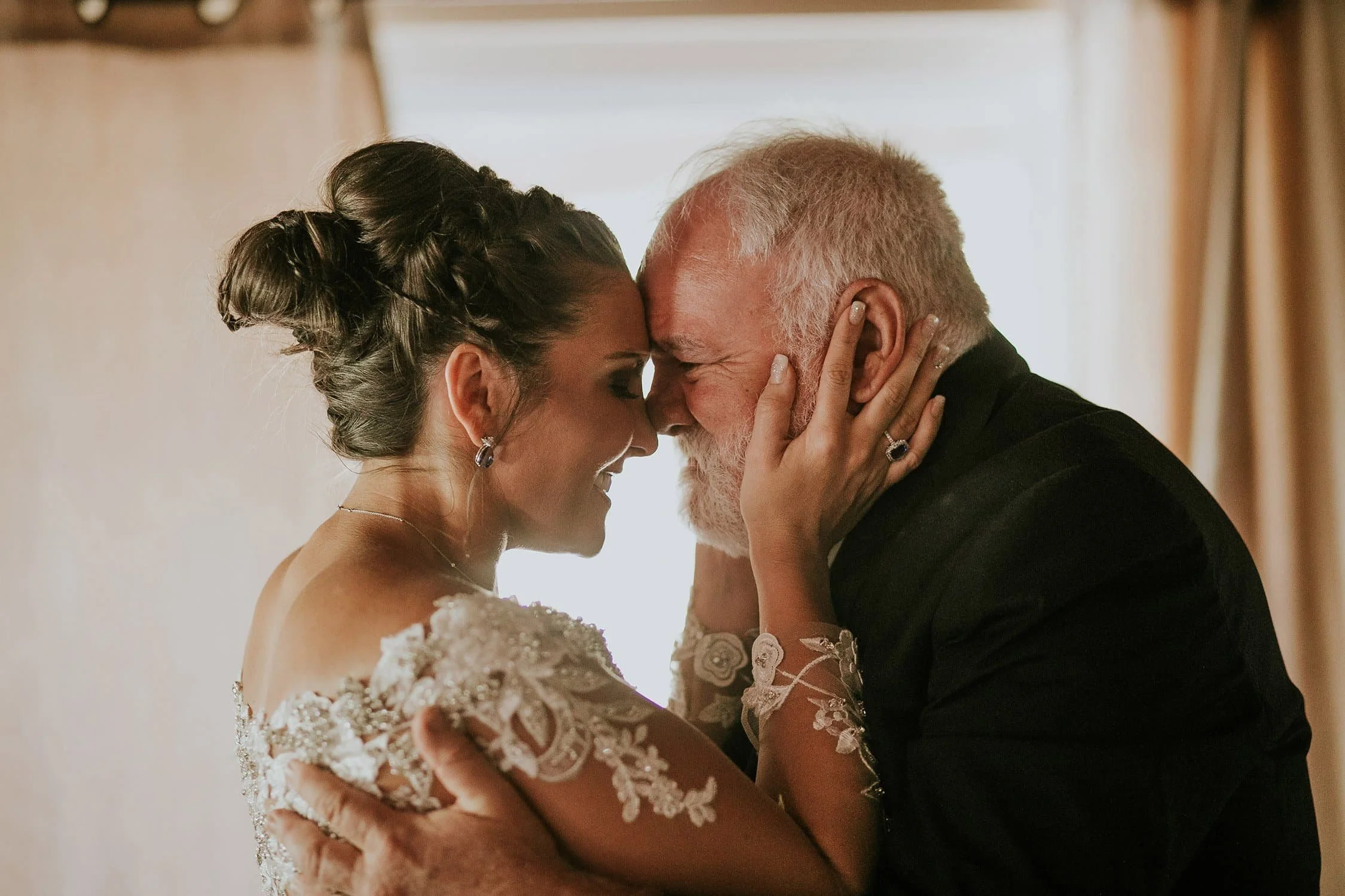 A bride and an elderly man, likely her father, shared a warm, intimate moment with their foreheads touching and eyes closed, smiling softly during a wedding celebration.