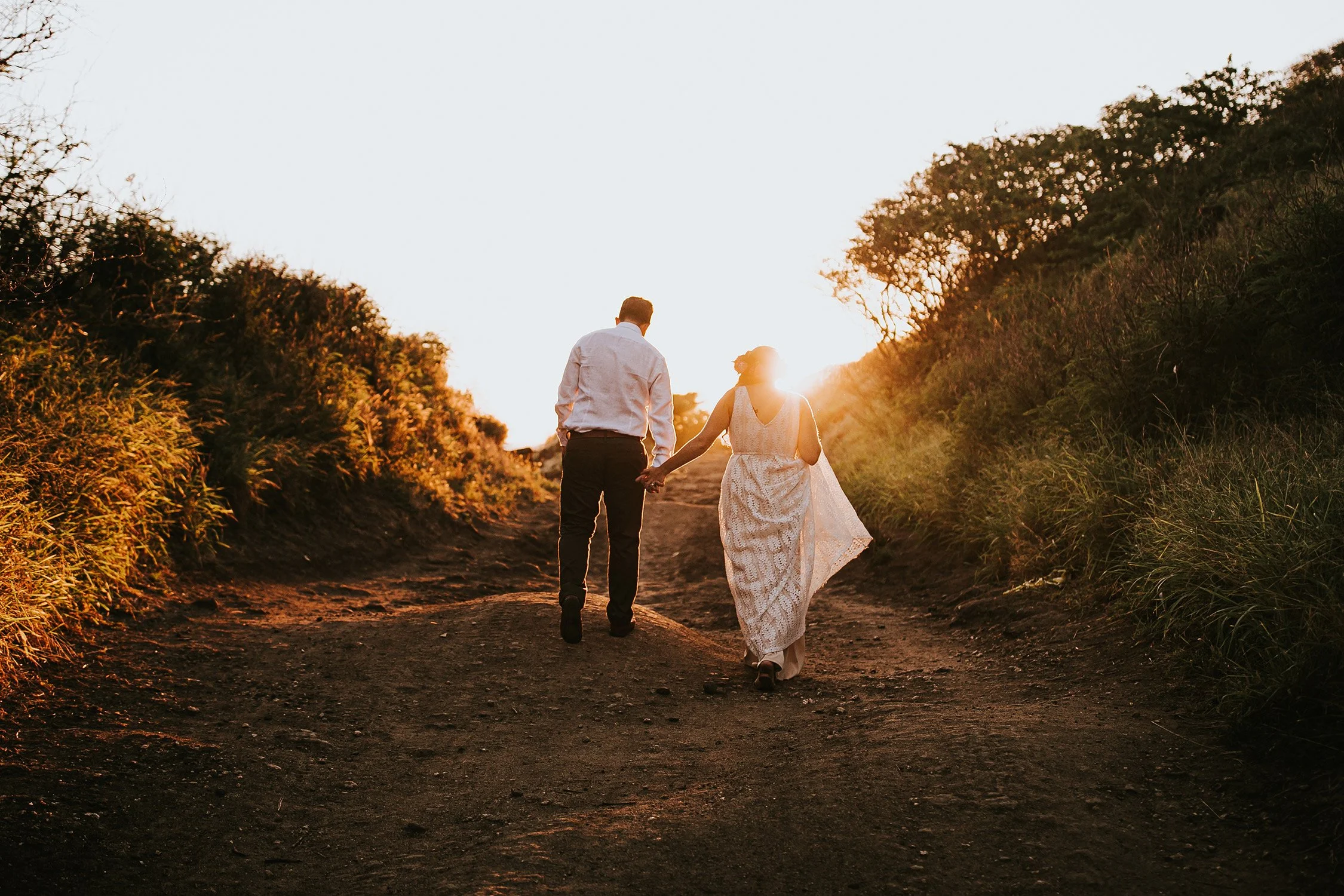 A couple holding hands and walking on a dirt path through a countryside landscape during sunset, with trees lining the path on both sides.