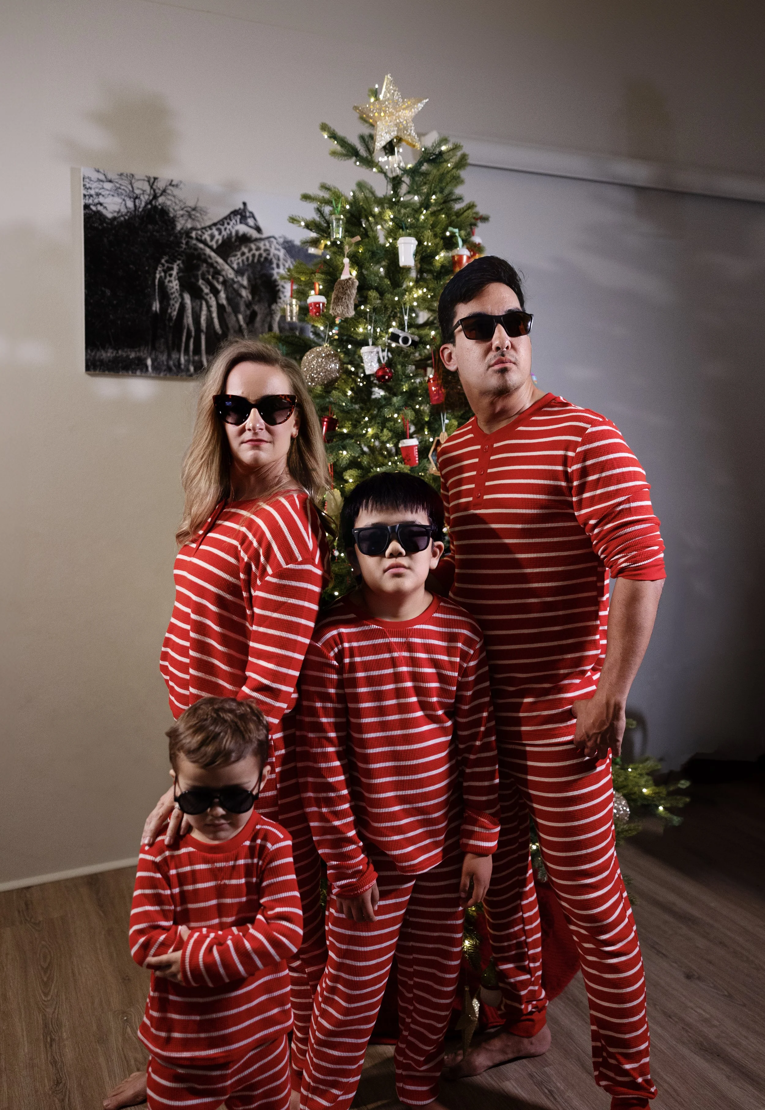 A family of five dressed in matching red and white striped pajamas, posing in front of a decorated Christmas tree, all wearing sunglasses.