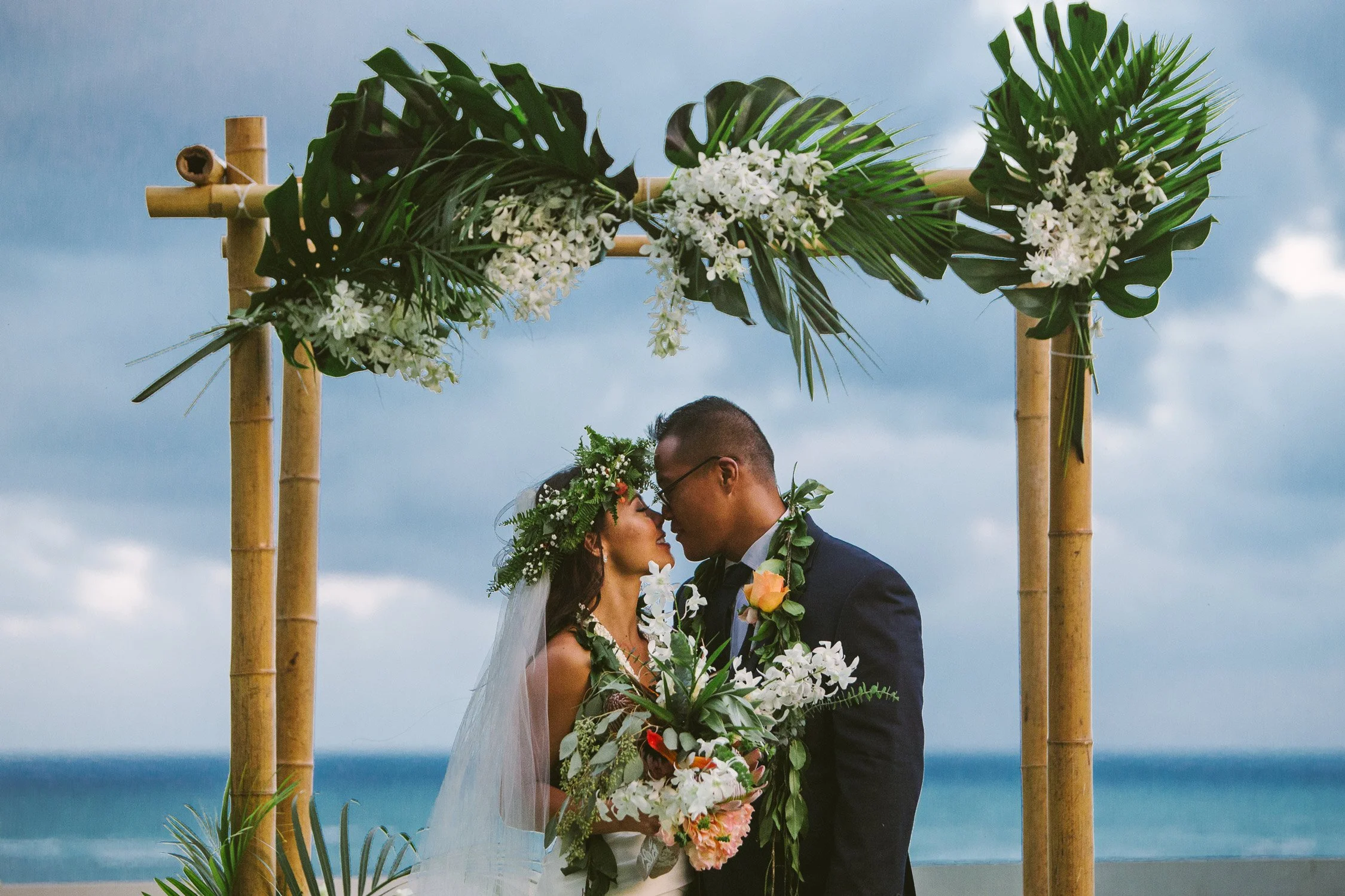 A couple getting married outdoors, standing under a bamboo arch decorated with tropical leaves and white flowers, with the ocean and cloudy sky in the background.