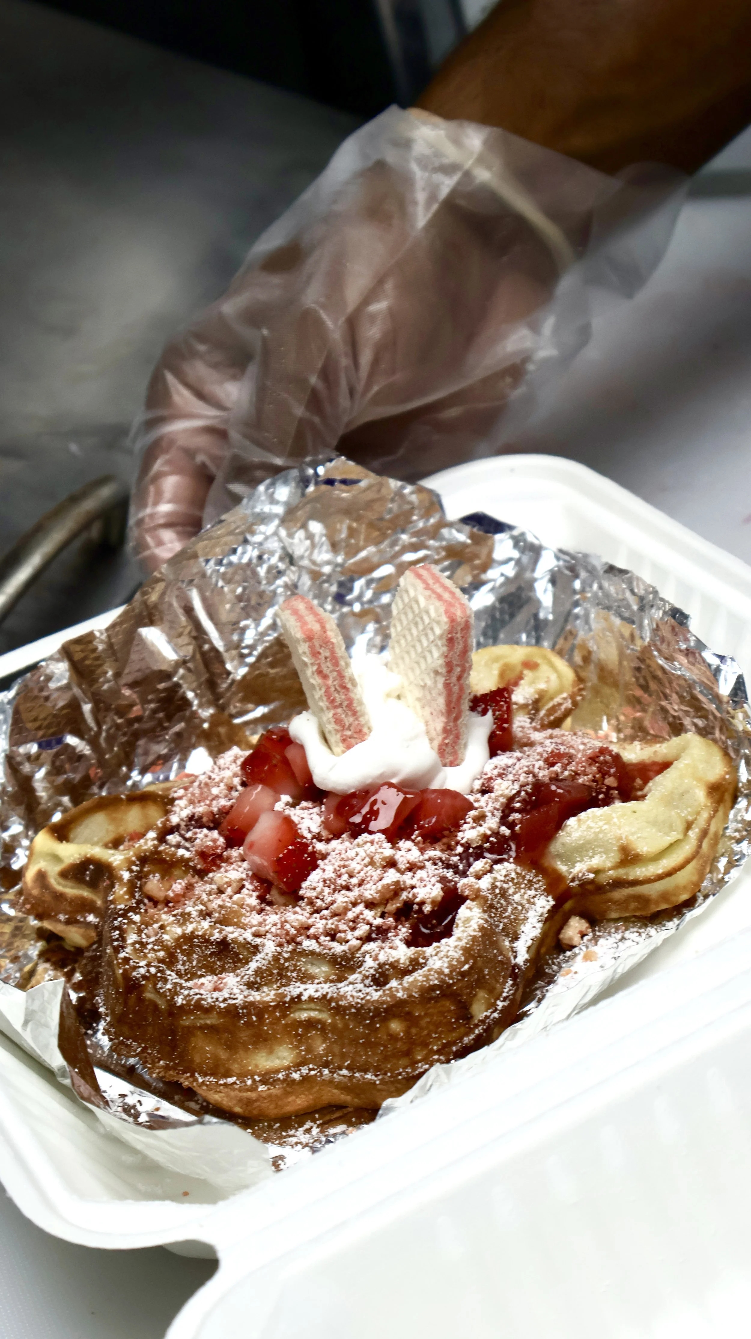 A dessert topped with strawberries, whipped cream, wafers, and caramel syrup in aluminum foil on a foam tray.