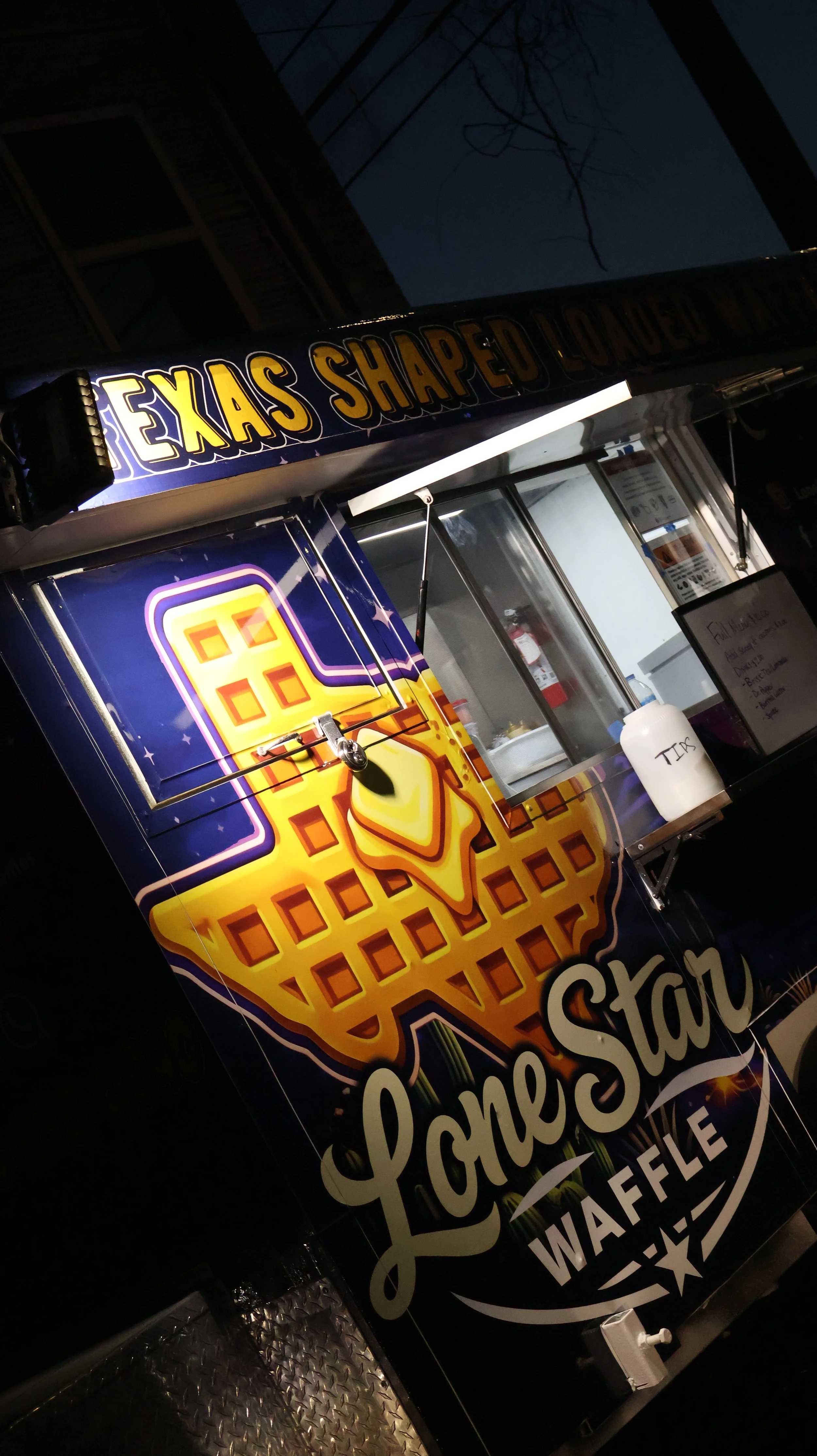 A colorful food truck with yellow waffle graphic and text that reads 'Lone Star' and 'Waffles'. The top sign says 'Texas Shaped Loaded'. The truck has a service window with a white container labeled 'TIPS' nearby, set against a dark night sky.