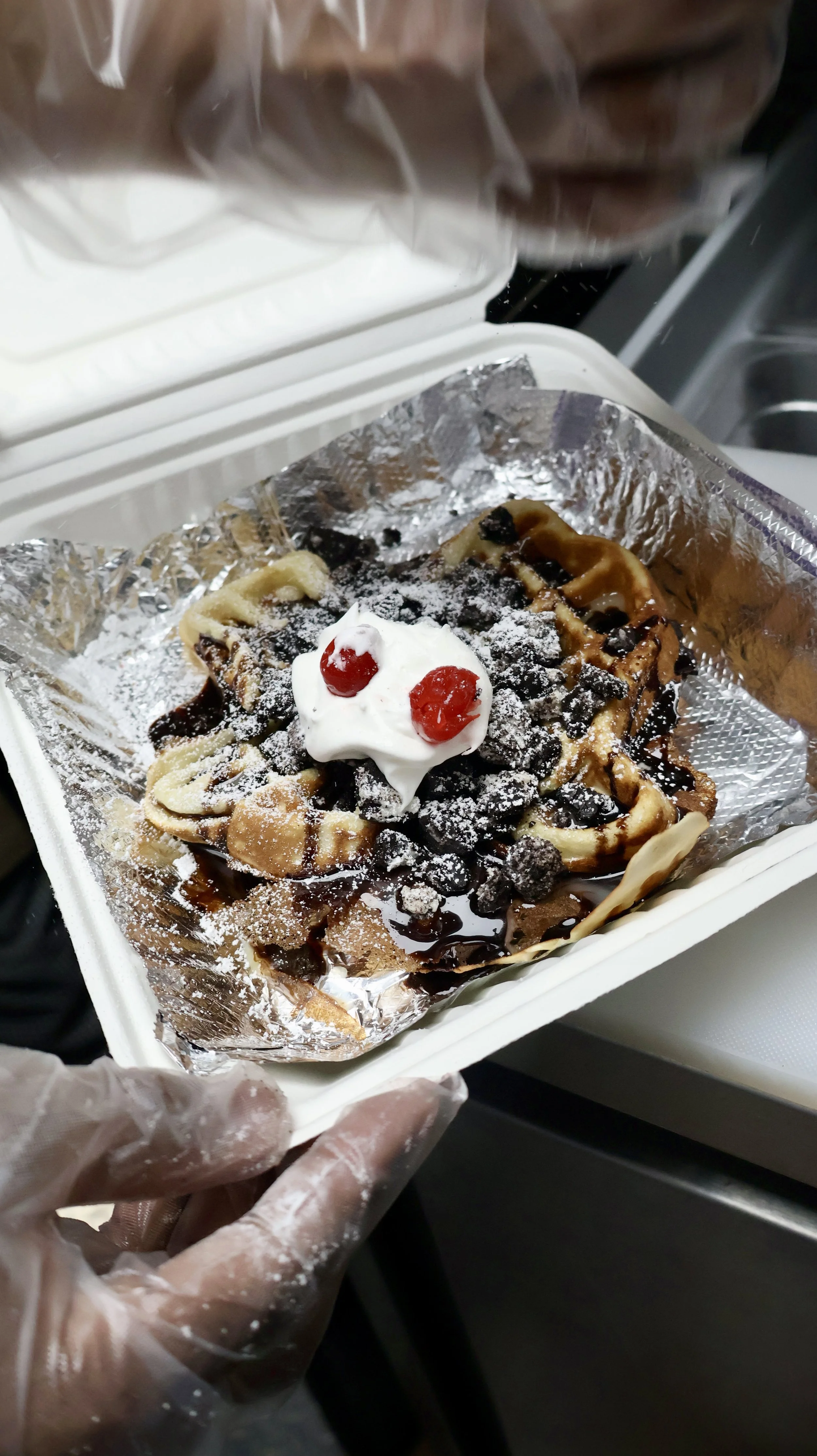 A waffle topped with whipped cream, maraschino cherries, chocolate sauce, chocolate chips, and powdered sugar, in a takeout container lined with foil.