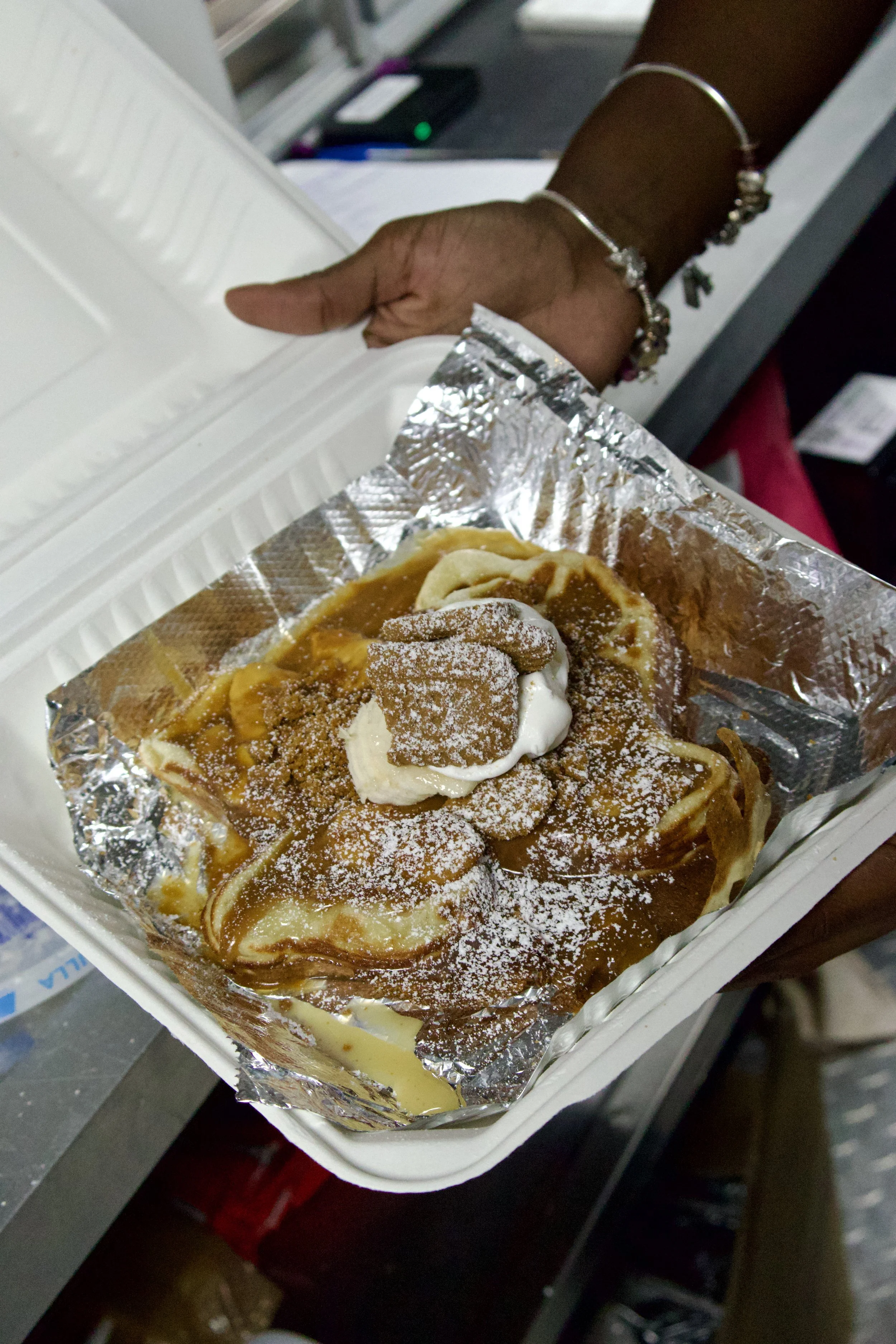 A hand holding a square of Belgian waffle topped with a dollop of whipped cream, caramel sauce, and a sprinkle of powdered sugar, with a burger box and a cash register in the background.
