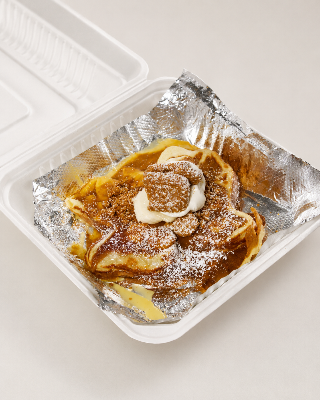 A serving of Mexican dessert, tres leches cake topped with whipped cream, caramel sauce, powdered sugar, and a cookie, inside a white plastic takeout container lined with aluminum foil.
