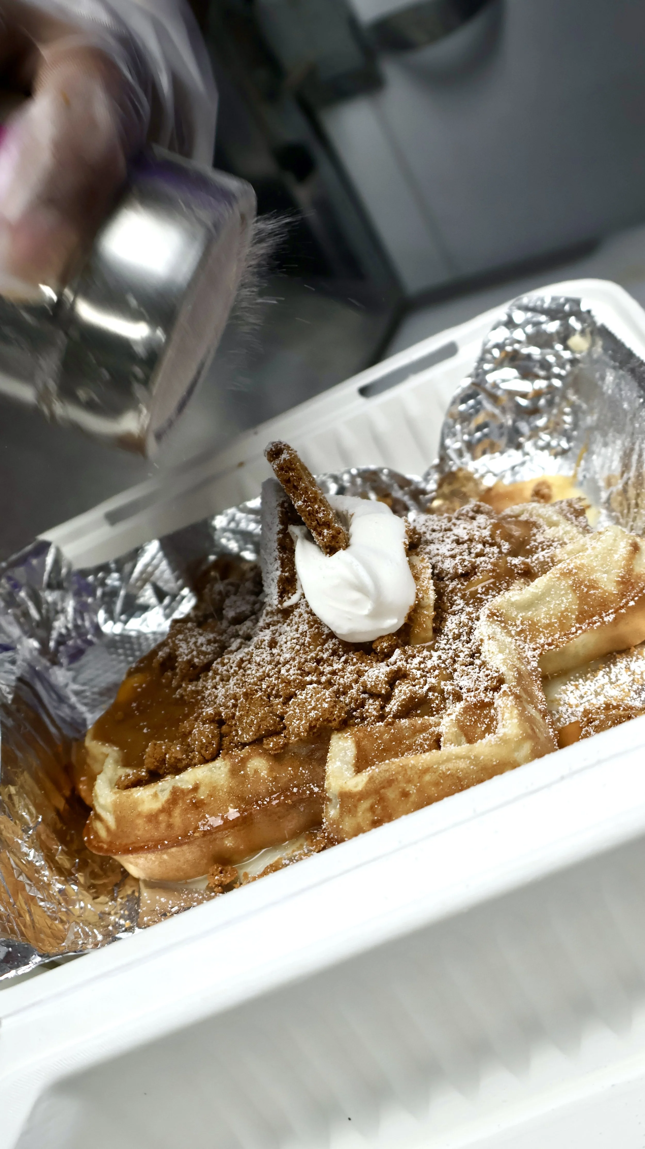 A waffle drizzled with caramel sauce, topped with whipped cream and a pretzel stick, all in a white takeout container.