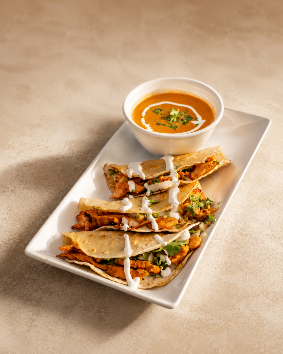 Plate of three chicken tacos with cilantro and sauce, served with a bowl of spicy tomato soup garnished with cream and cilantro.