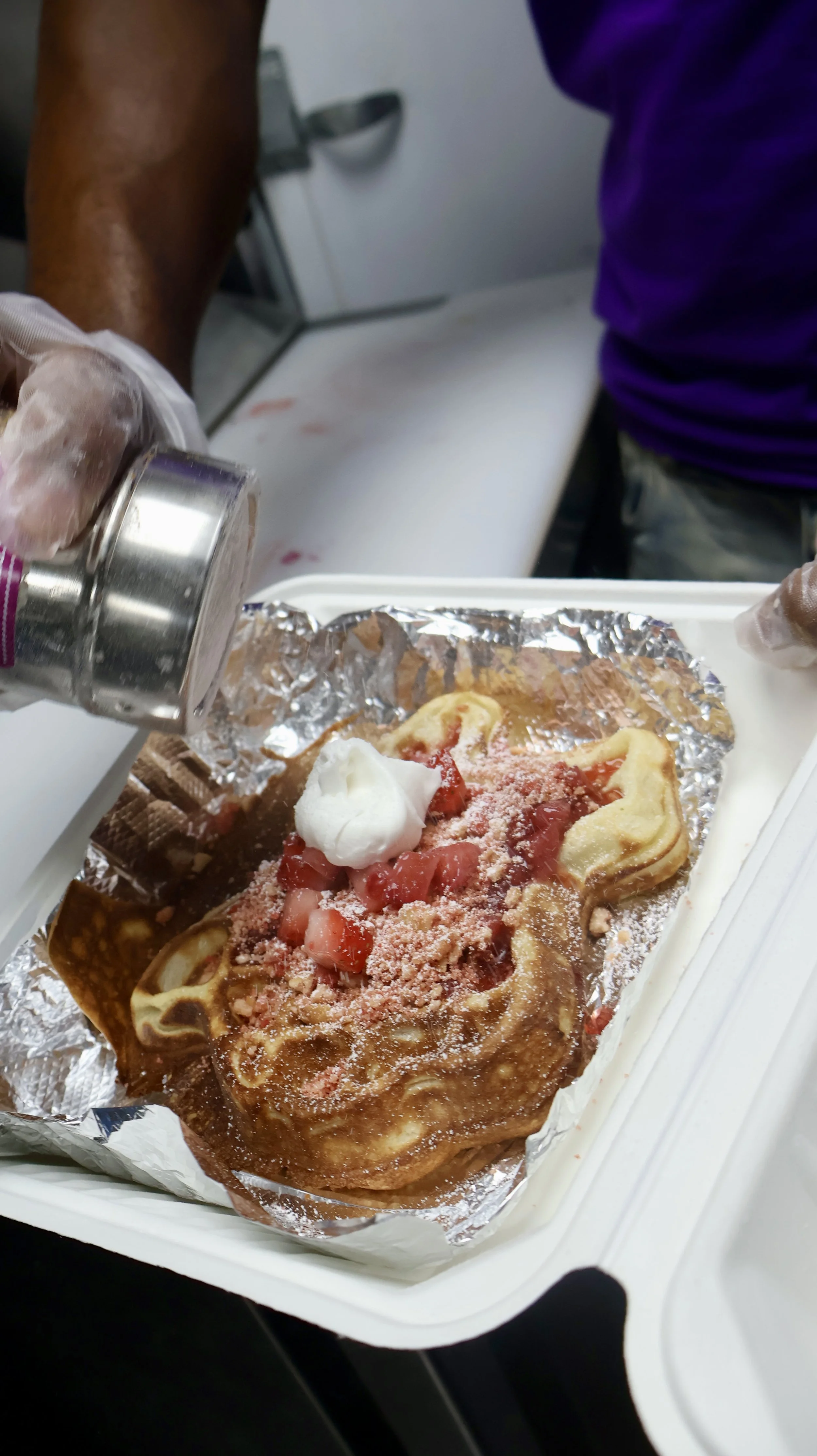 A person wearing gloves is pouring a topping onto a large slice of pizza in a food service setting. The pizza appears to have strawberries, whipped cream, and pink powder on top.
