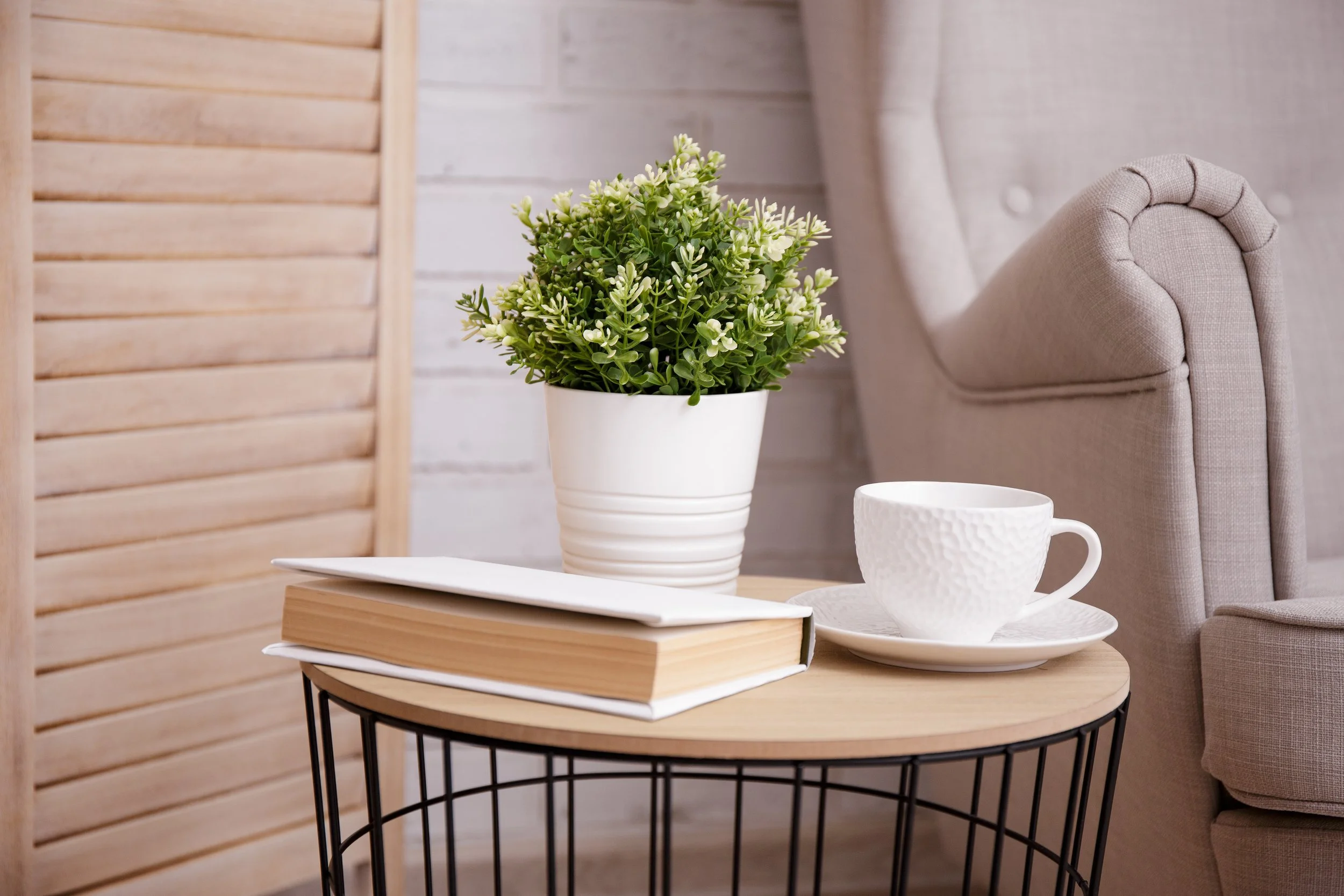 A beige upholstered armchair, a small wooden table with a black wire frame, a white ceramic cup and saucer, two stacked books with beige covers, a white vase with green and white artificial plant, and a wooden room divider with horizontal slats.