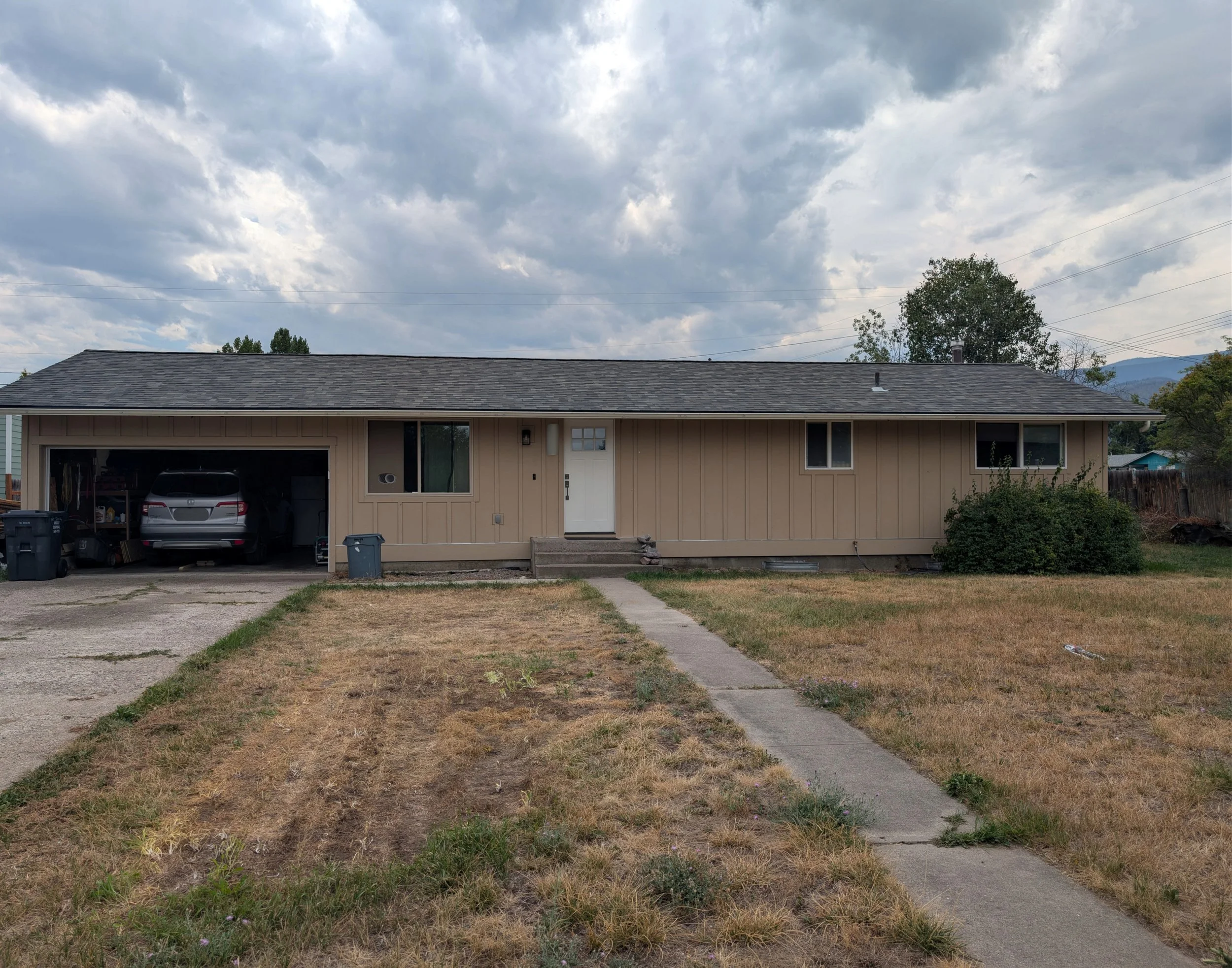 A single-story house with a gray shingle roof, beige siding, a front door, and windows. A driveway with a car parked inside and trash bins at the front. The yard has a concrete sidewalk and patchy grass. The sky is cloudy with some blue visible and trees and mountains in the background.